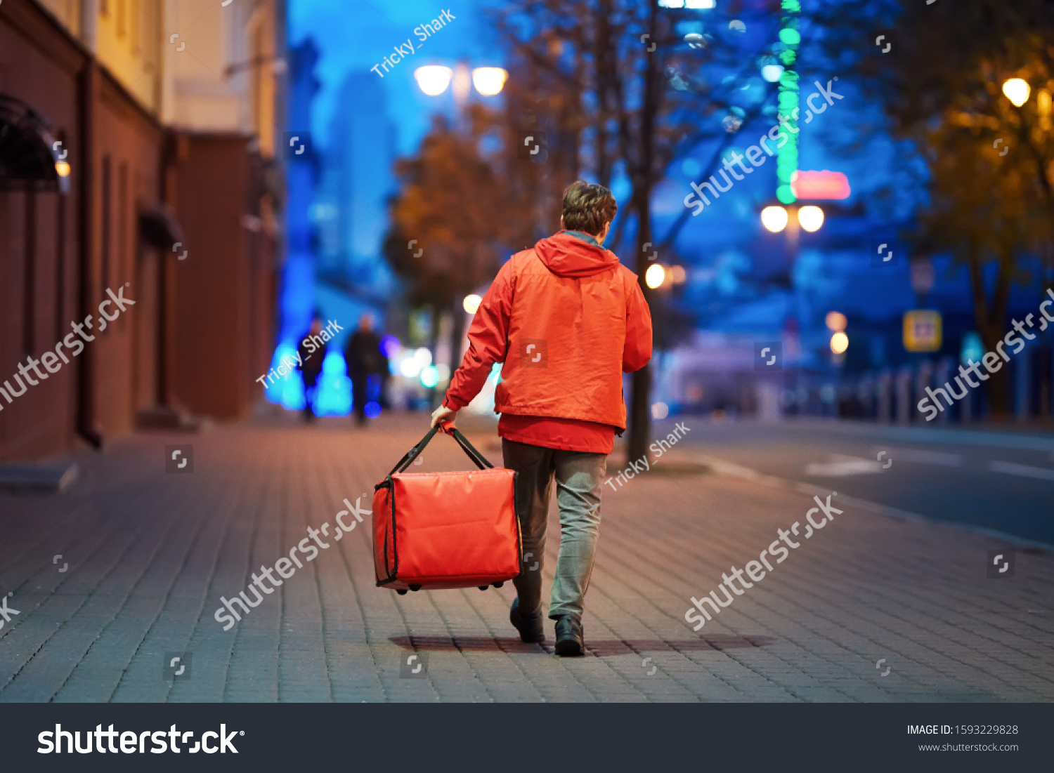 Young deliveryboy walking with red thermal bag on night city street. Man of delivery service in hurry to deliver an order. Delivery service goes to give the order quickly to the client at night #1593229828