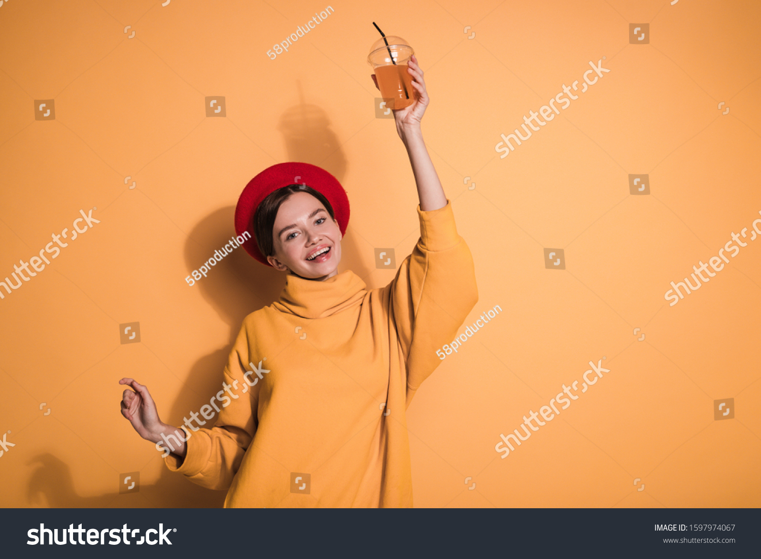 Cheerful playful young woman posing on camera alone. Hold drink in one hand. Look straight and smile. Wear red beret and stylish sweater. Isolated over yellow background #1597974067