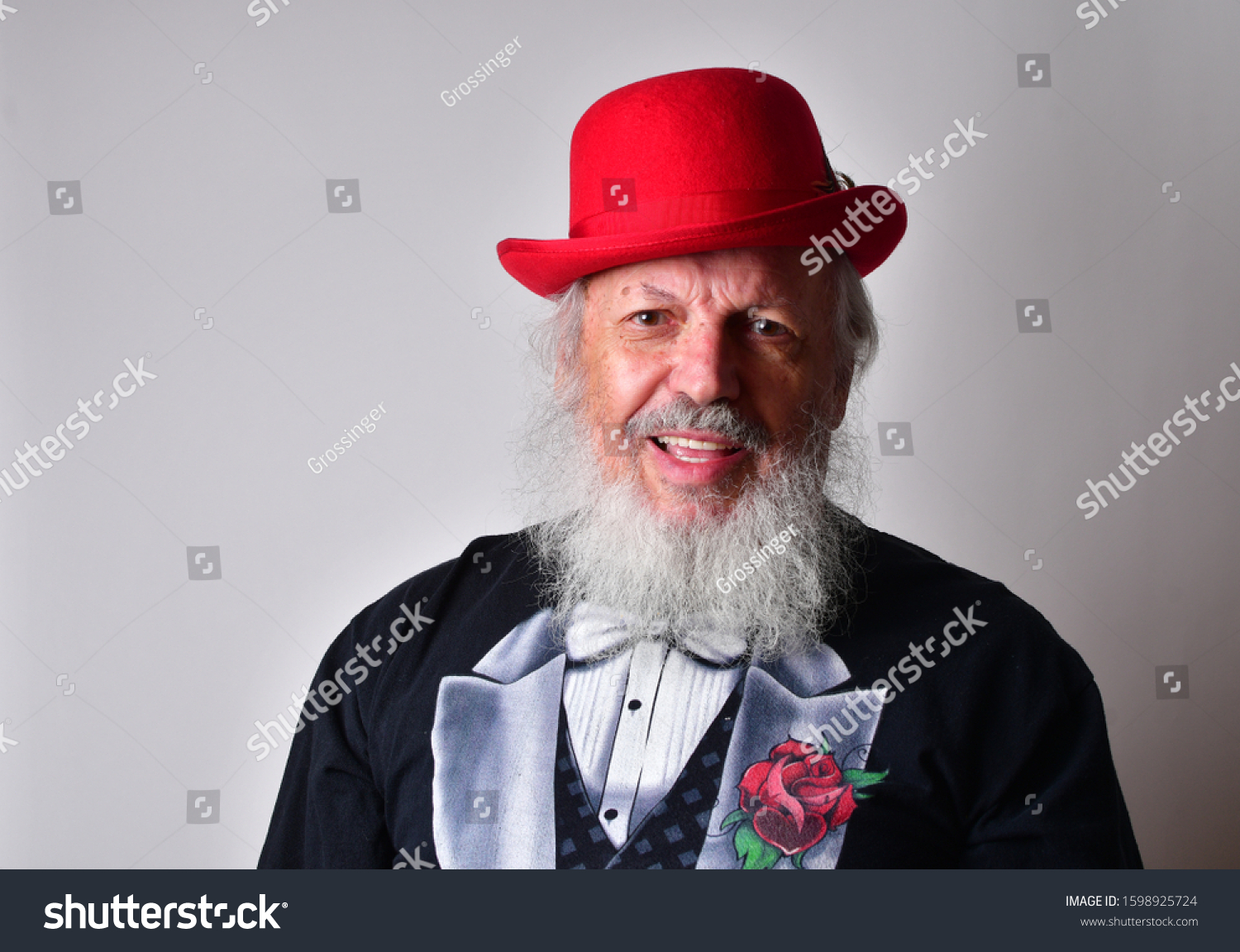 Happy old man in a fake tuxedo wearing a red bowler hat and making facial expressions.
Old Caucasian with a long white beard, faux tuxedo and red bowler hat making  faces. #1598925724