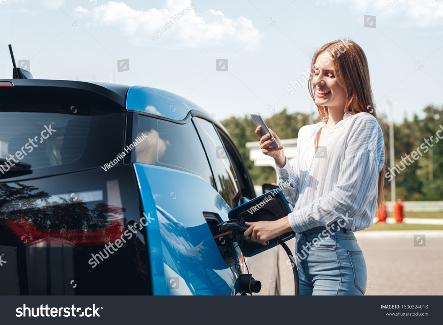 Young beautiful woman traveling by electric car having stop at charging station standing plugging cable browsing internet on smartphone smiling joyful while charing #1600324018