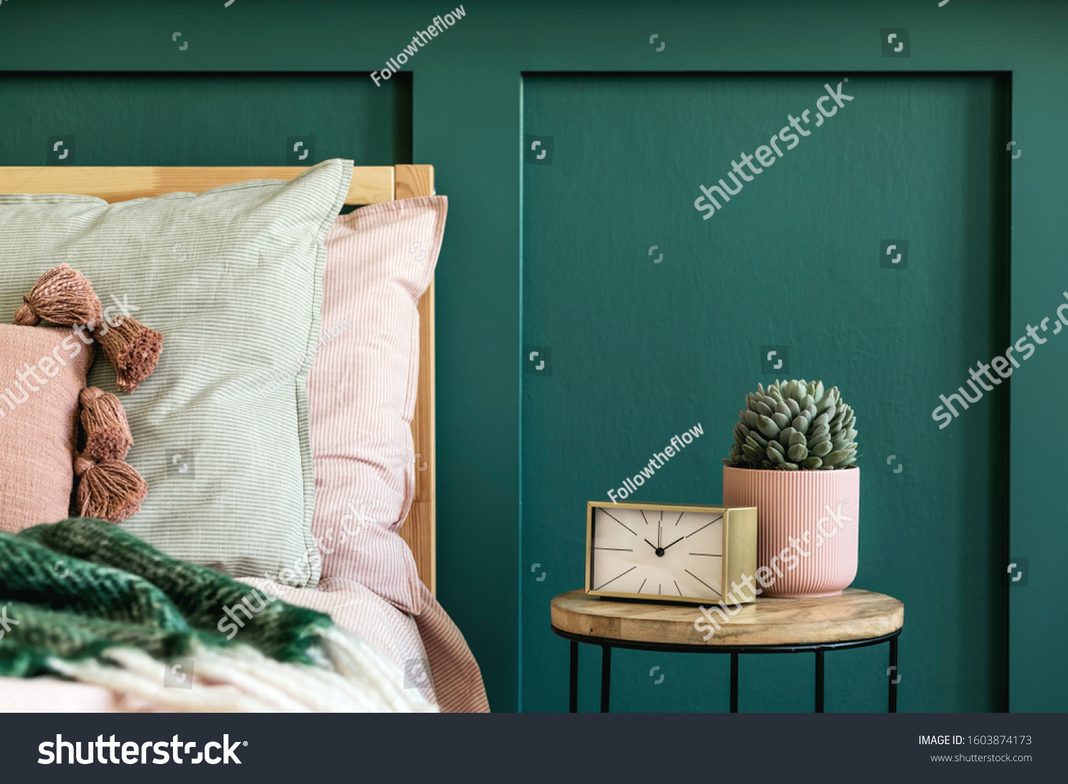 Stylish bedroom interior with design coffee table, plant, gold clock and elegant personal accessories. Beautiful bed sheets, blanket and pillows. Template. Modern home staging. Wall panelling. Details #1603874173