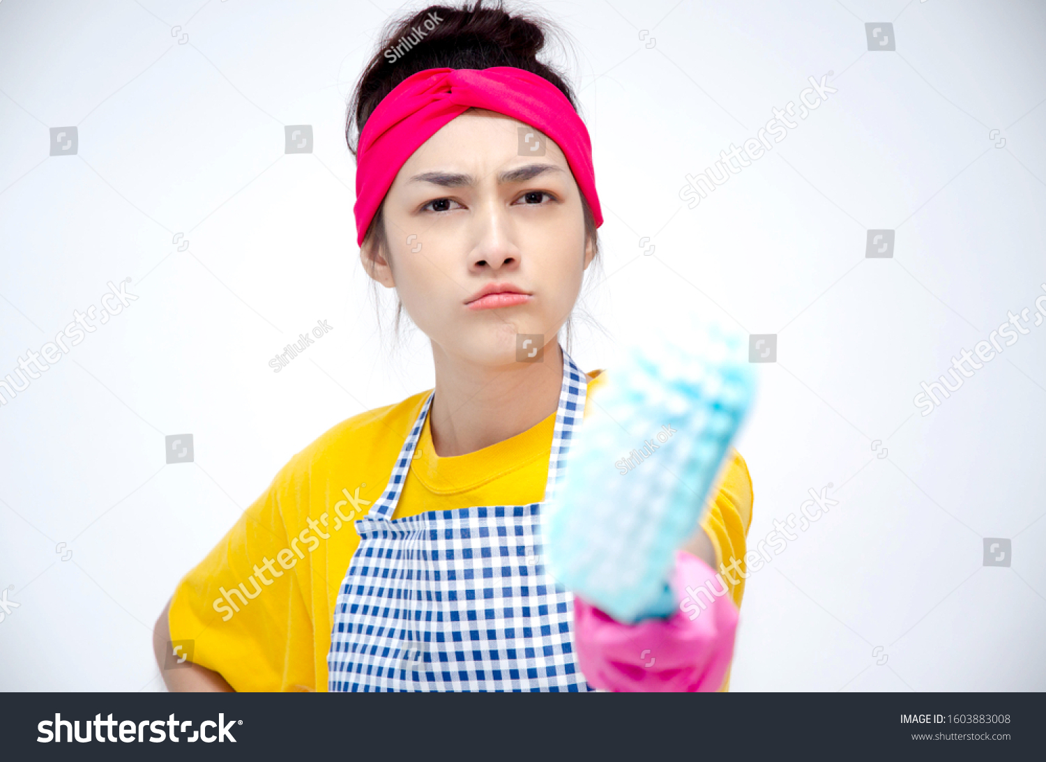 The housewife wears yellow clothes, wears an apron, wears pink gloves, stands for the index finger and makes stressful faces. Copy area. #1603883008