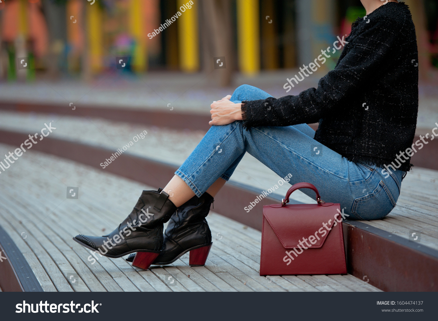 Fashionable young woman in high heel black cowboy boots, blue jeans, black tweed jacket and burgundy handbag on the city street. Street style. #1604474137