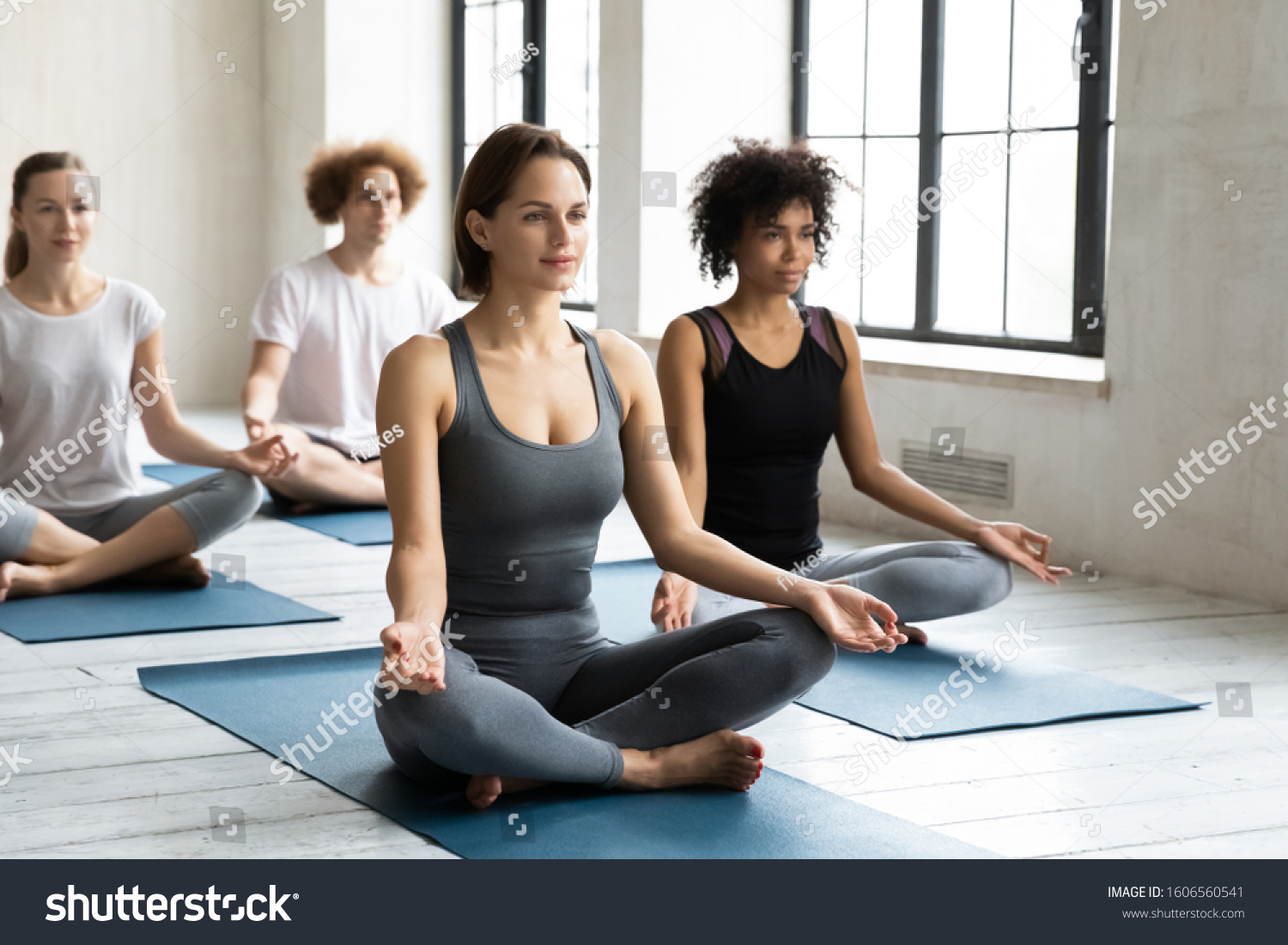 Diverse young people sitting in Easy Seat pose on mats, practicing yoga at group lesson, meditating in Sukhasana exercise, working out in modern yoga studio, stress relief and wellness #1606560541