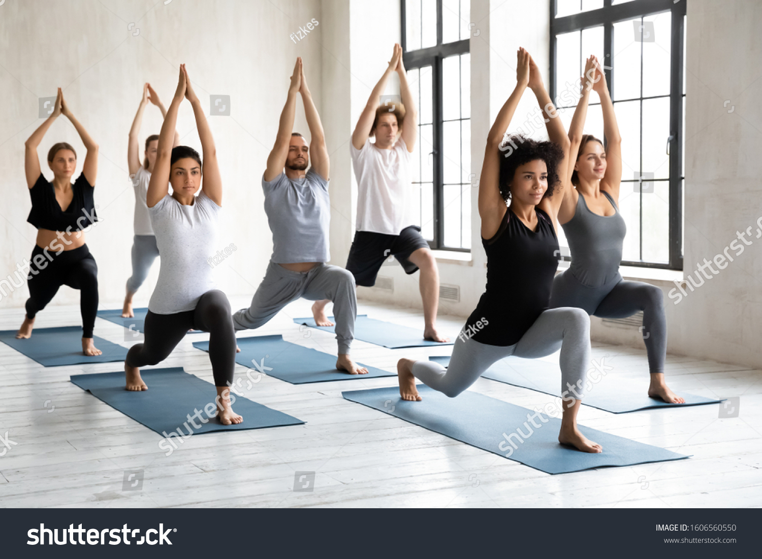 Young diverse young people with African American instructor doing Warrior one exercise, practicing yoga at group lesson, standing in Virabhadrasana pose, working out in modern yoga studio #1606560550