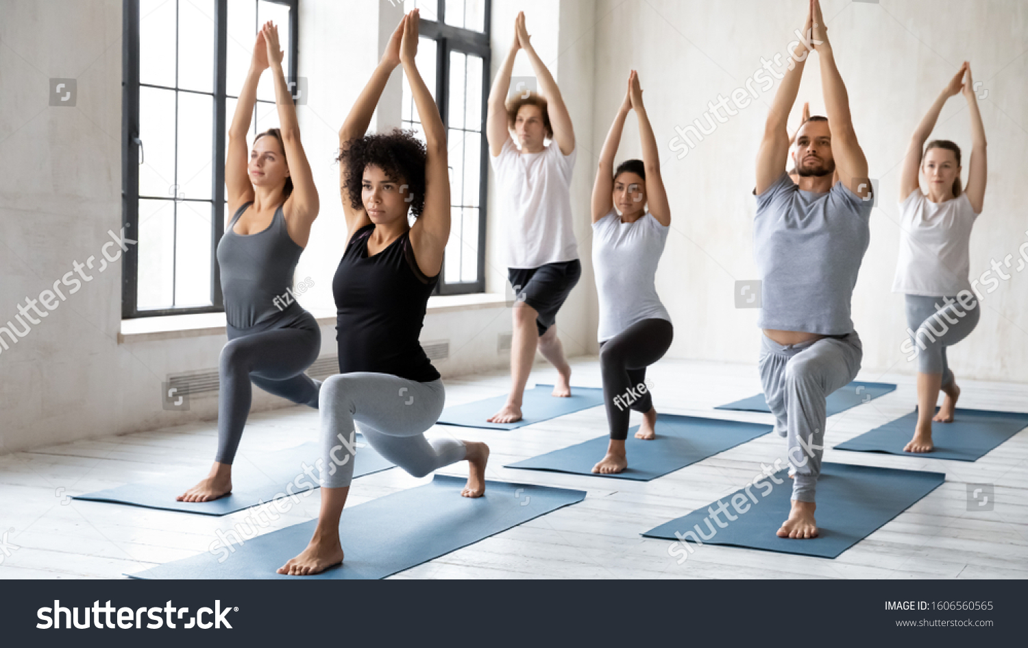 Diverse young sporty people doing Warrior one exercise at group lesson, practicing yoga in modern fitness center, standing in Virabhadrasana pose, working out with African American female instructor #1606560565