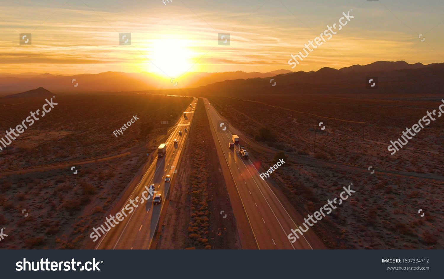 DRONE, SUN FLARE: Scenic shot of 18 wheeler trucks and cars crossing Mojave desert at dusk. Golden evening sun rays shine on the traffic moving up and down the straight freeway in rural California. #1607334712