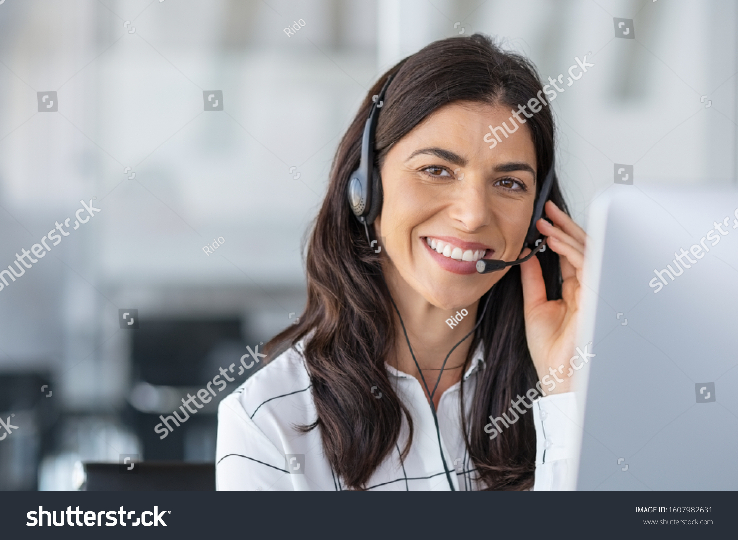 Call center agent with headset working on support hotline in modern office with copy space. Portrait of mature positive agent in conversation with customer over headset looking at camera. #1607982631