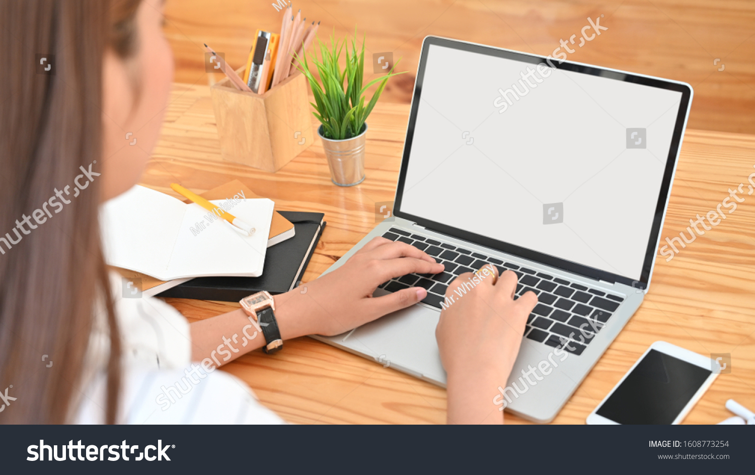 Photo views from back of young woman while using/typing on white blank screen laptop including pencil holder, black blank screen smartphone, potted plant, pencil, notes, earphone on wooden desk. #1608773254