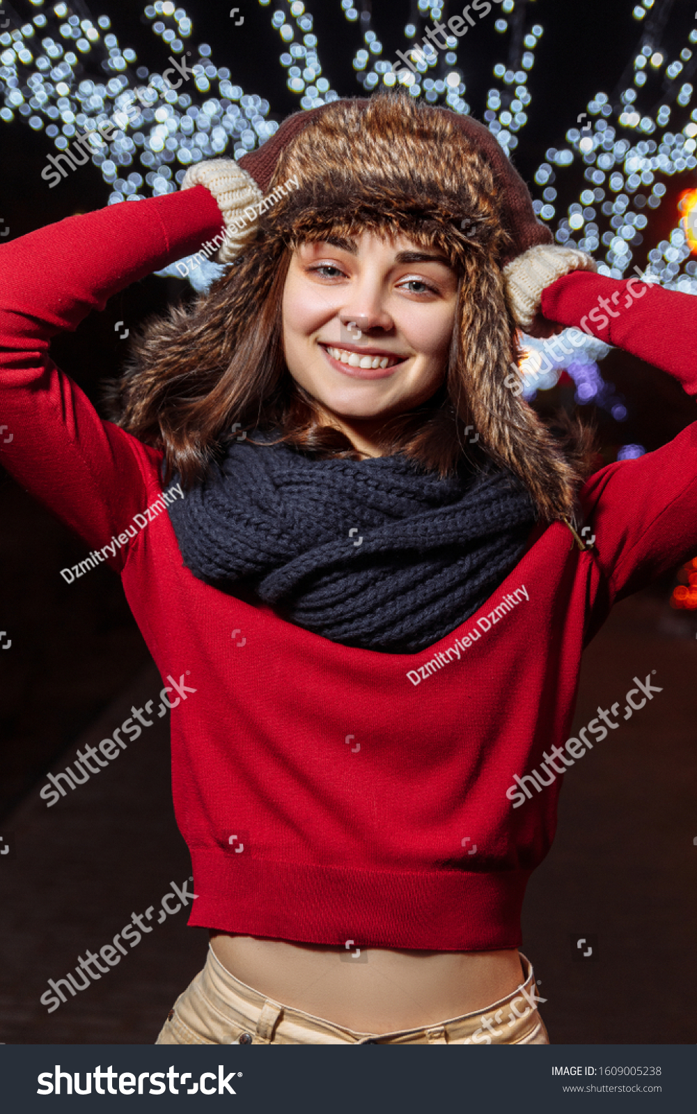 Young charming woman in red sweater and warm hat standing on street with illuminated sparkling Christmas decorations in winter and looking at camera #1609005238