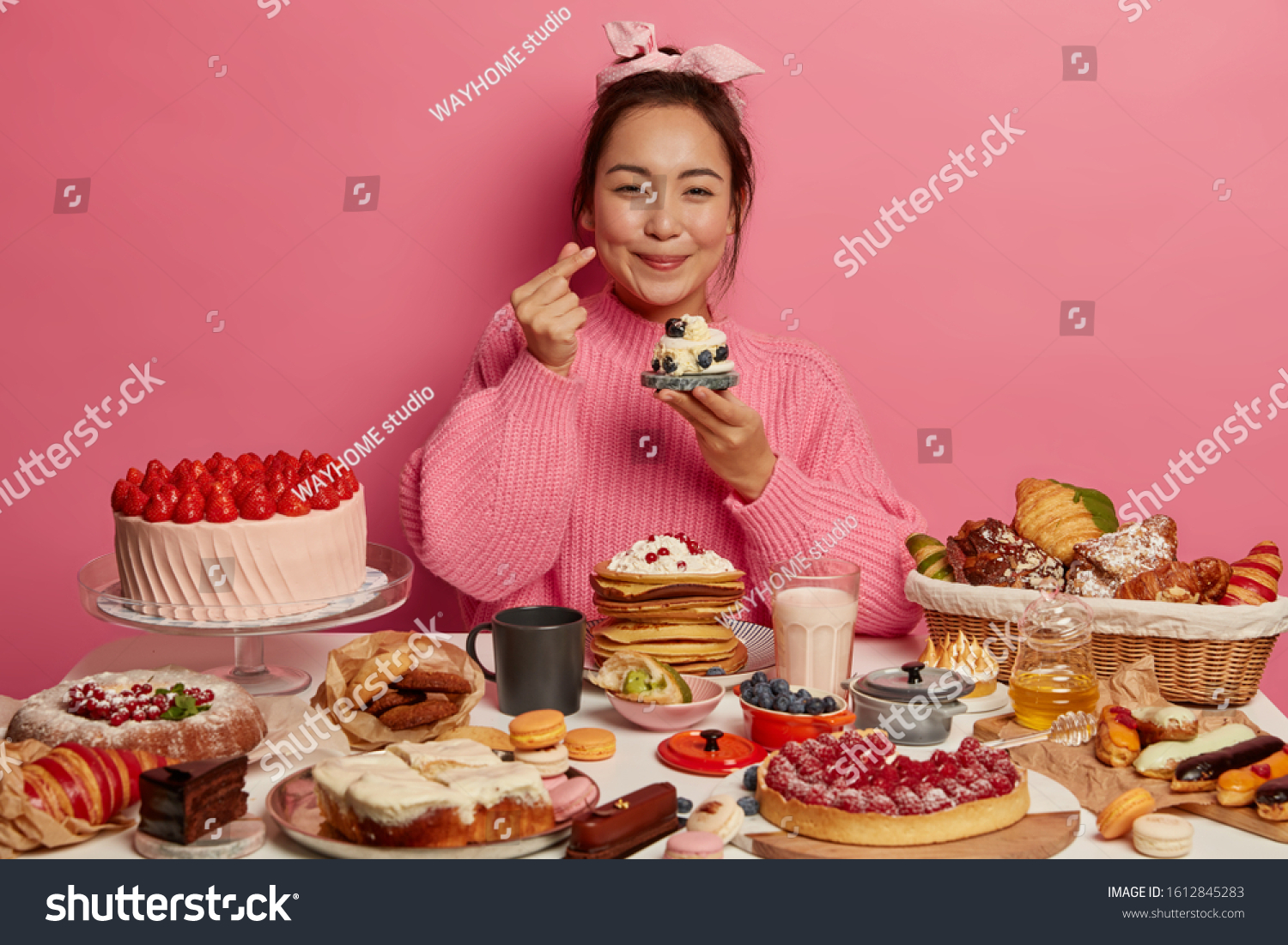 Shot of adorable woman makes korean like sign, expresses affection, holds small delicious cupcake, celebrates birthday, isolated over rosy wall, made many tasty desserts. Cheat meal concept. #1612845283