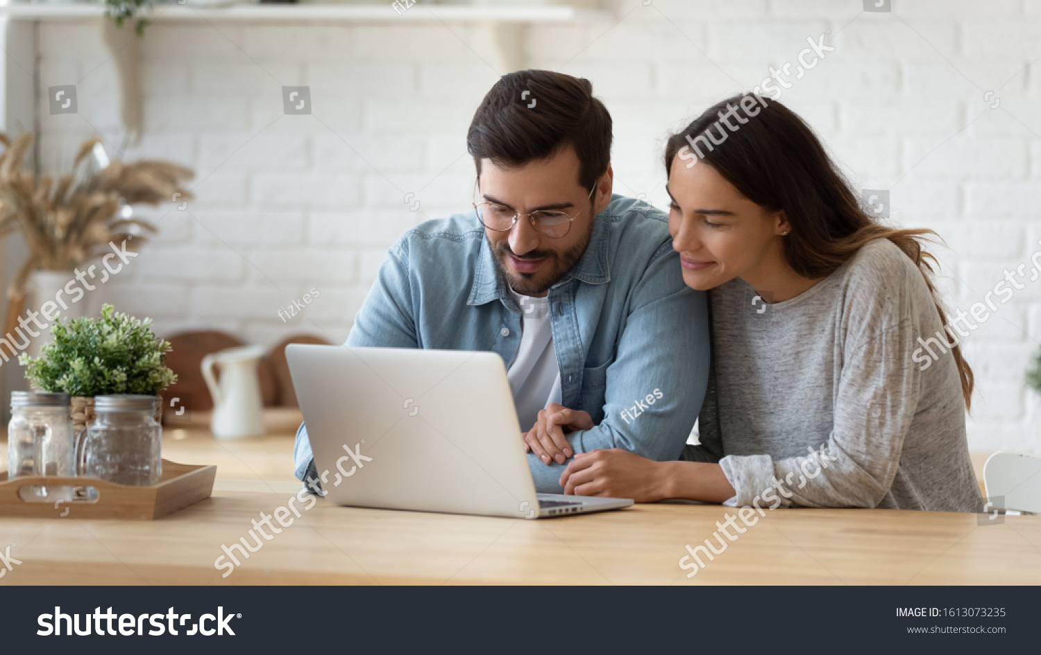 Pleasant family couple sitting at big wooden table in modern kitchen, looking at laptop screen. Happy young mixed race married spouse web surfing, making purchases online or booking flight tickets. #1613073235