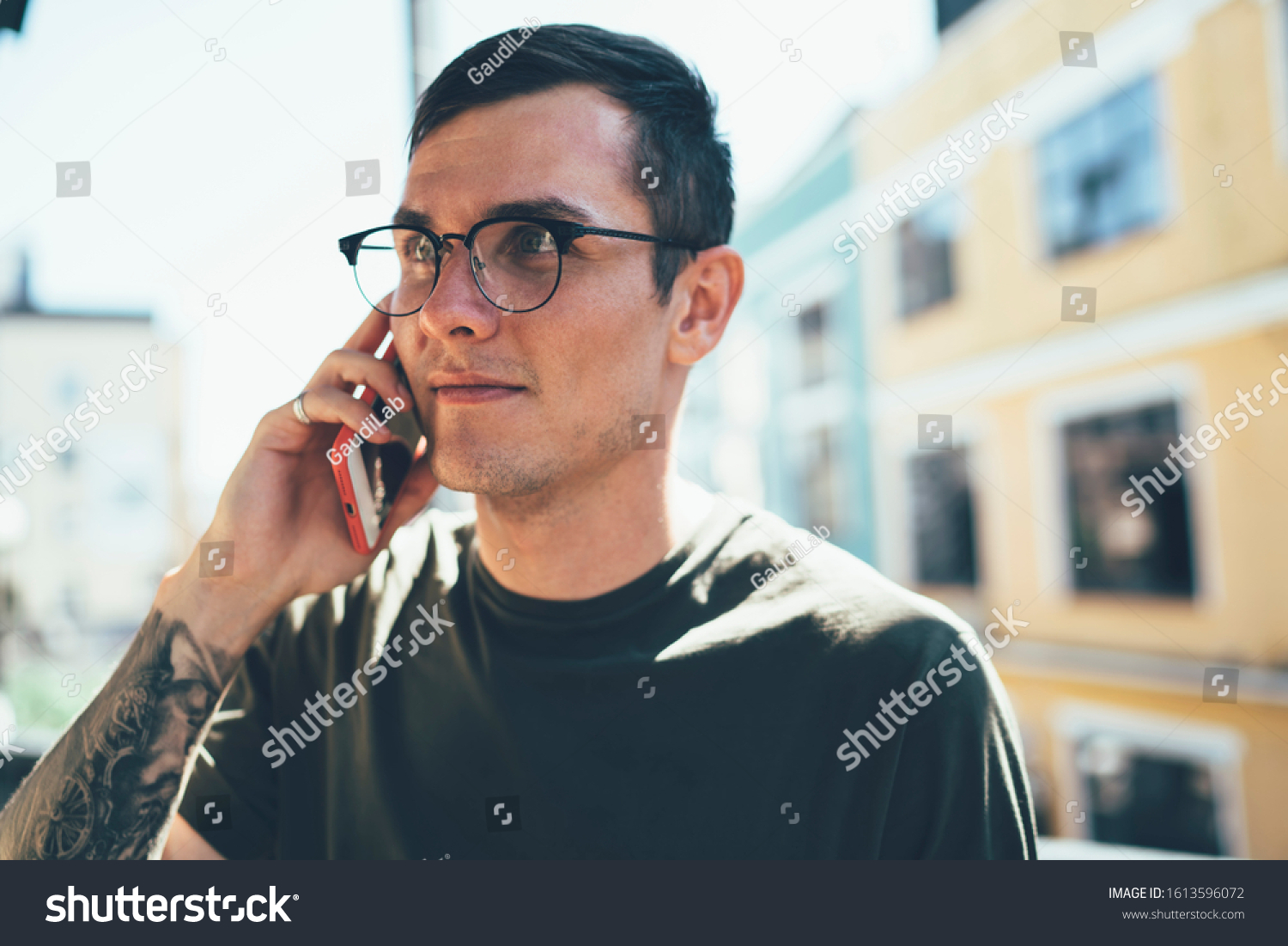 Concentrated young cool male in casual wear and glasses with tattoo using smartphone for discussion with coworker collaborative project listening ideas #1613596072