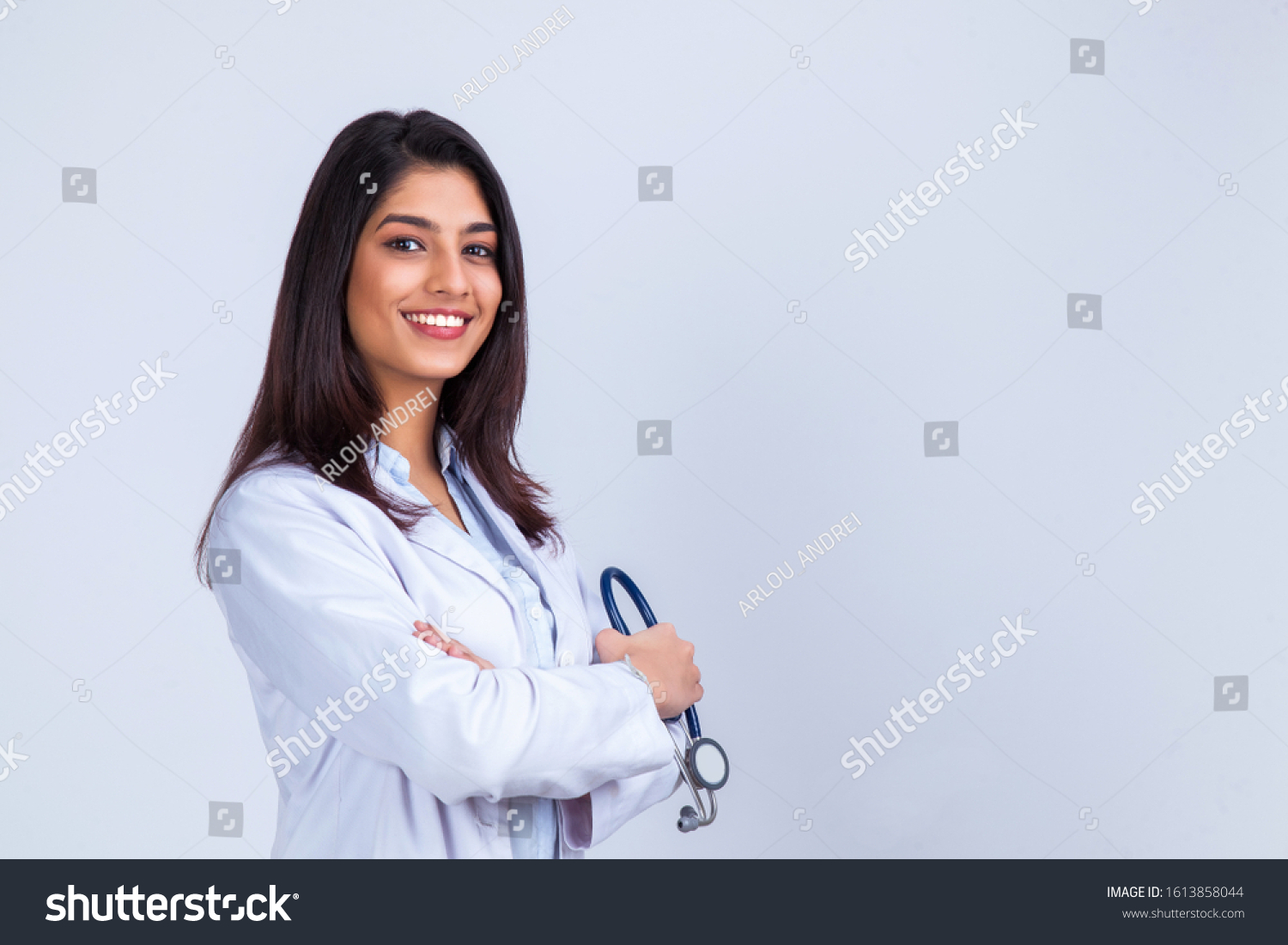 Medical concept of Indian beautiful female doctor in white coat with stethoscope, waist up. Medical student. Woman hospital worker looking at camera and smiling, studio, gray background #1613858044