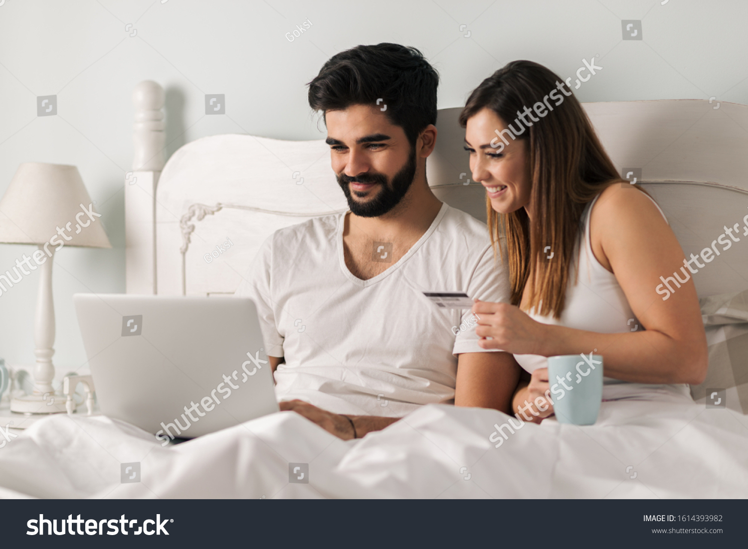 Smiling couple using laptop and credit card for e-commerce while sitting in bed #1614393982
