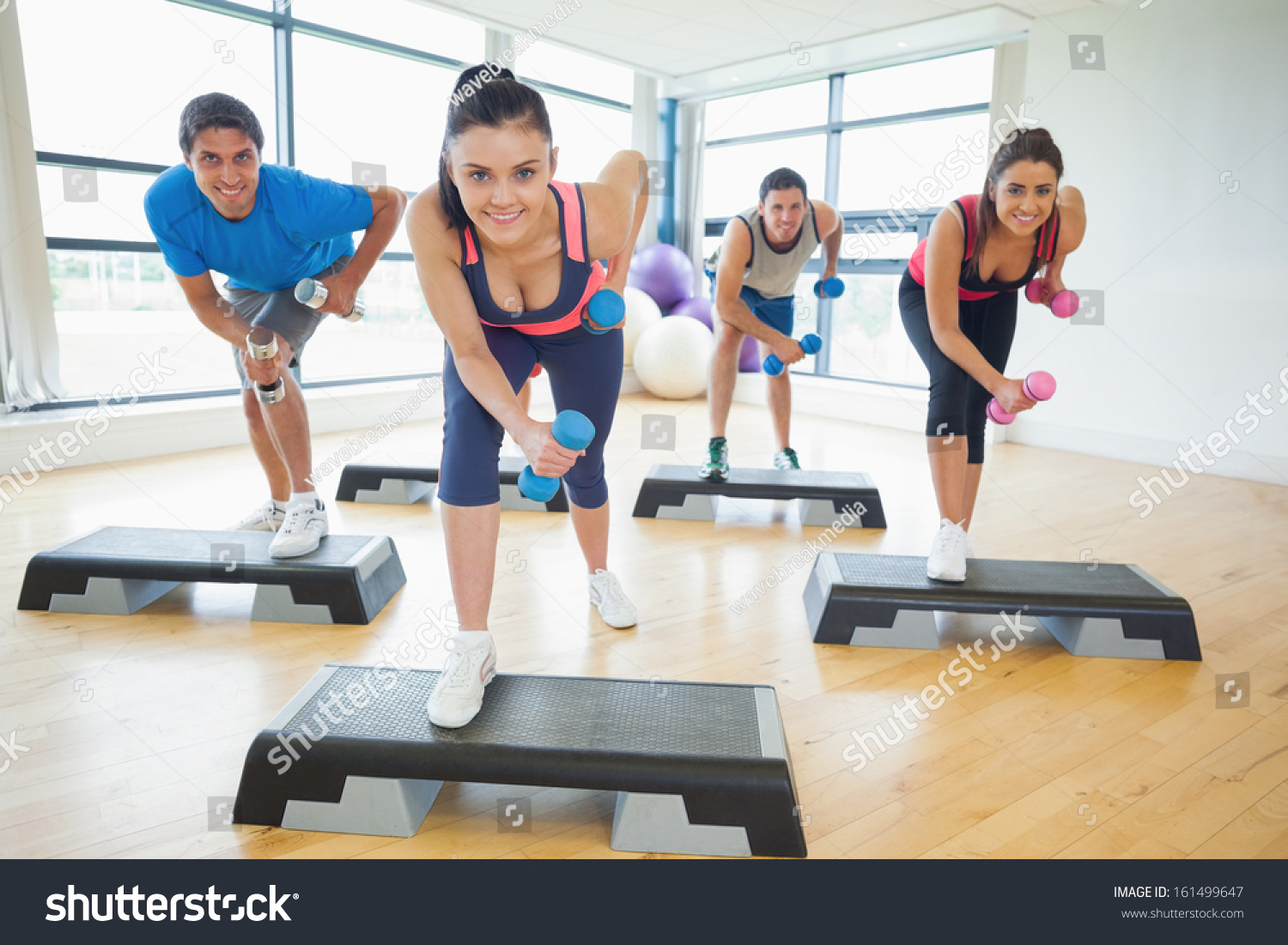 Full length of instructor with fitness class performing step aerobics exercise with dumbbells in a gym #161499647