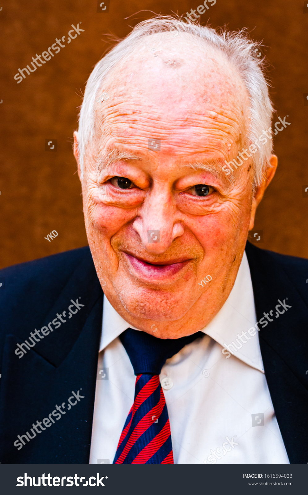 Portrait of happy white elderly man with dark suit, white shirt and striped blue red tie smiling on bright brown background #1616594023