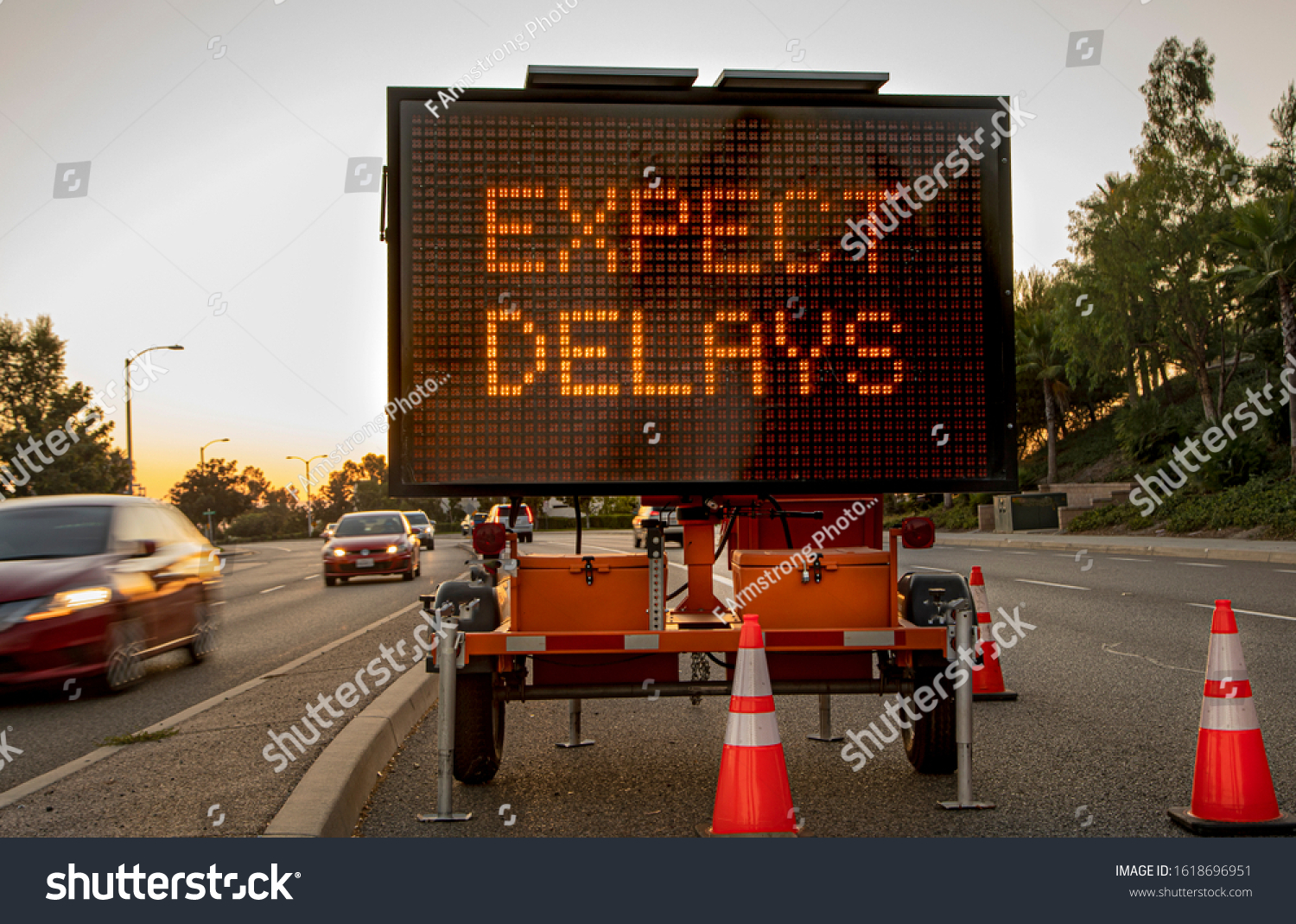 Mobile Electronic Traffic Sign stating “expect Delays” taken at sunset with traffic blurred driving past the sign and traffic cones #1618696951