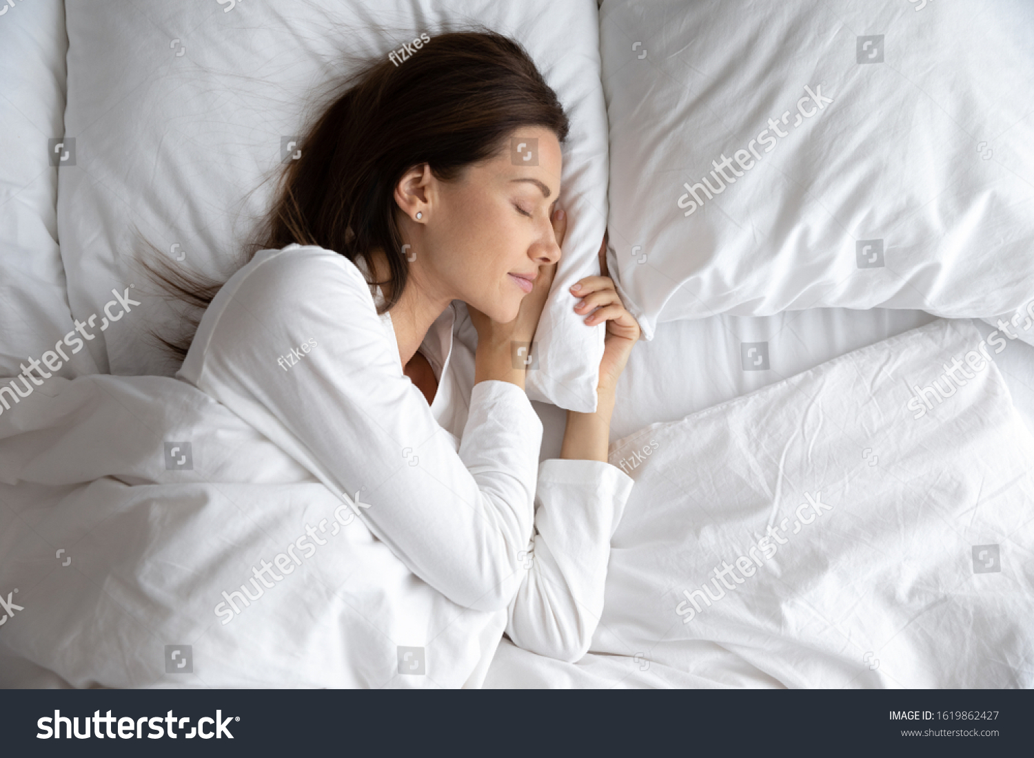 Serene beautiful woman sleeping with hand under cheek on soft pillow, young female wearing white pajamas resting under warm blanket in comfortable bed in bedroom or in hotel, enjoying sweet dreams #1619862427