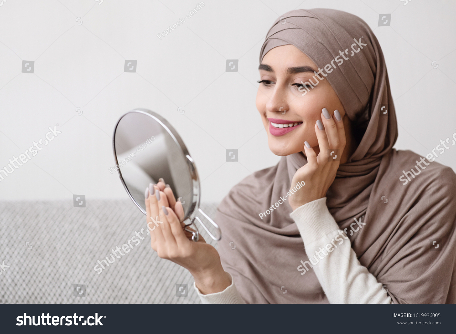 Beauty routine. Attractive muslim girl in hijab holding mirror and looking at her face with perfect soft skin, sitting on sofa at home, free space #1619936005