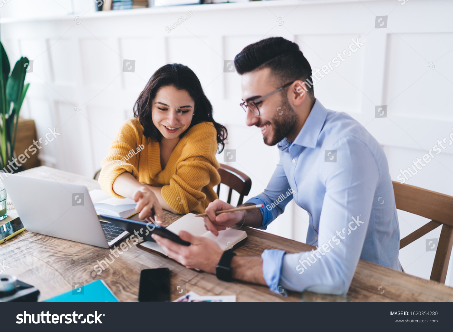 Glad couple wearing casual clothes writing notes in textbook while browsing laptop and tablet and sitting at table at home #1620354280