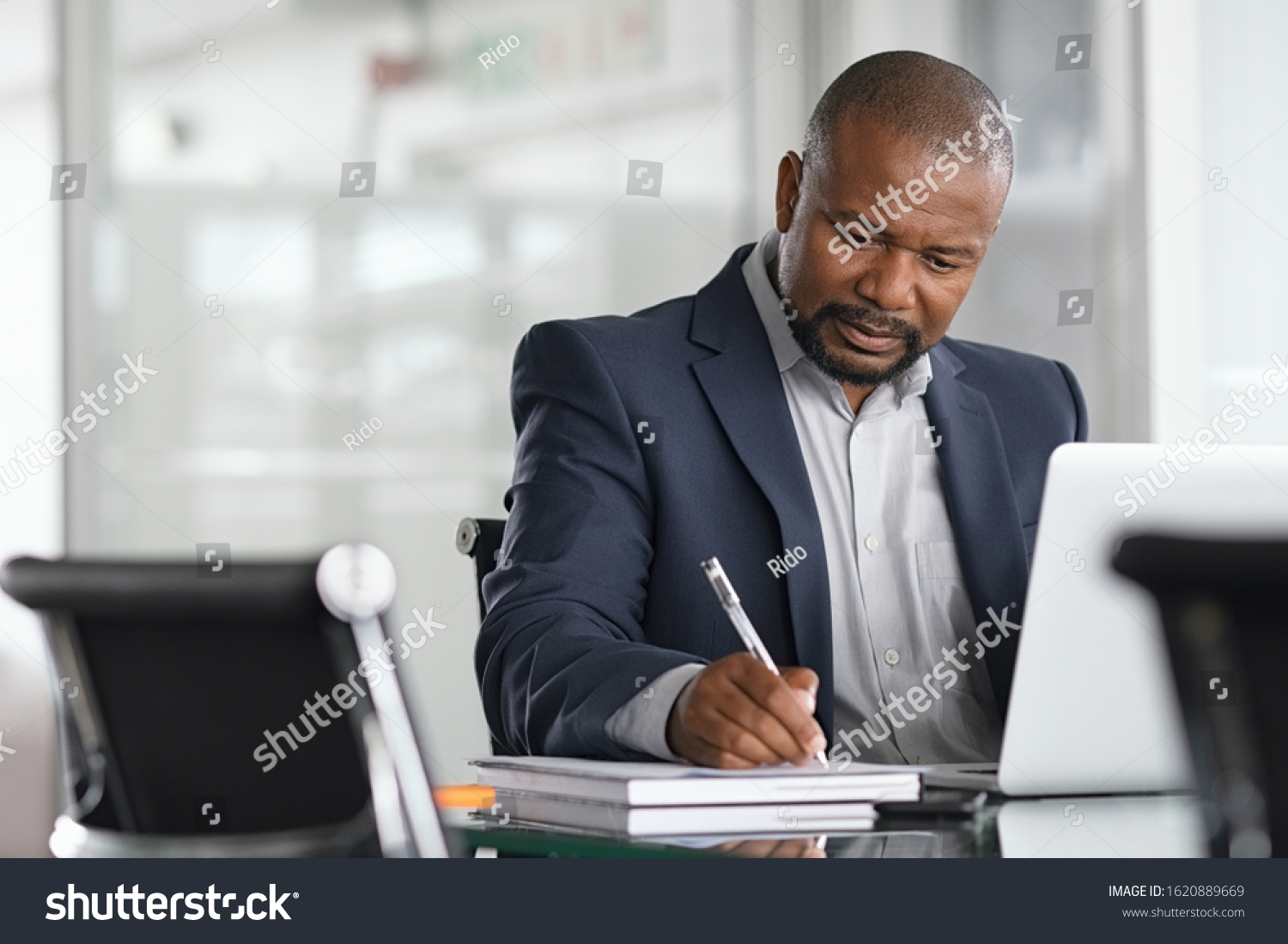 African serious businessman writing notes and using laptop. Mature business man writing his strategy in modern office. Focused black entrepreneur sitting at desk in modern office while working. #1620889669