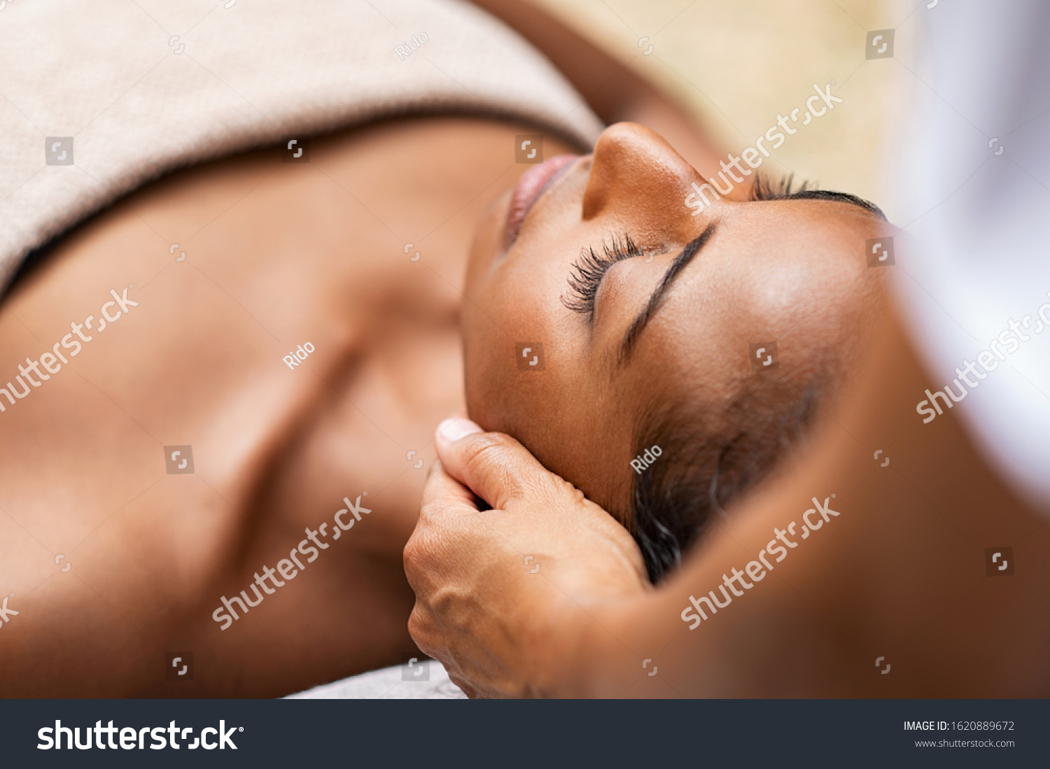 Beautiful african woman getting face massage in beauty spa. Black girl with closed eyes relaxing in outdoor spa while getting head massage. Serene woman relaxing outdoor in a beauty center. #1620889672