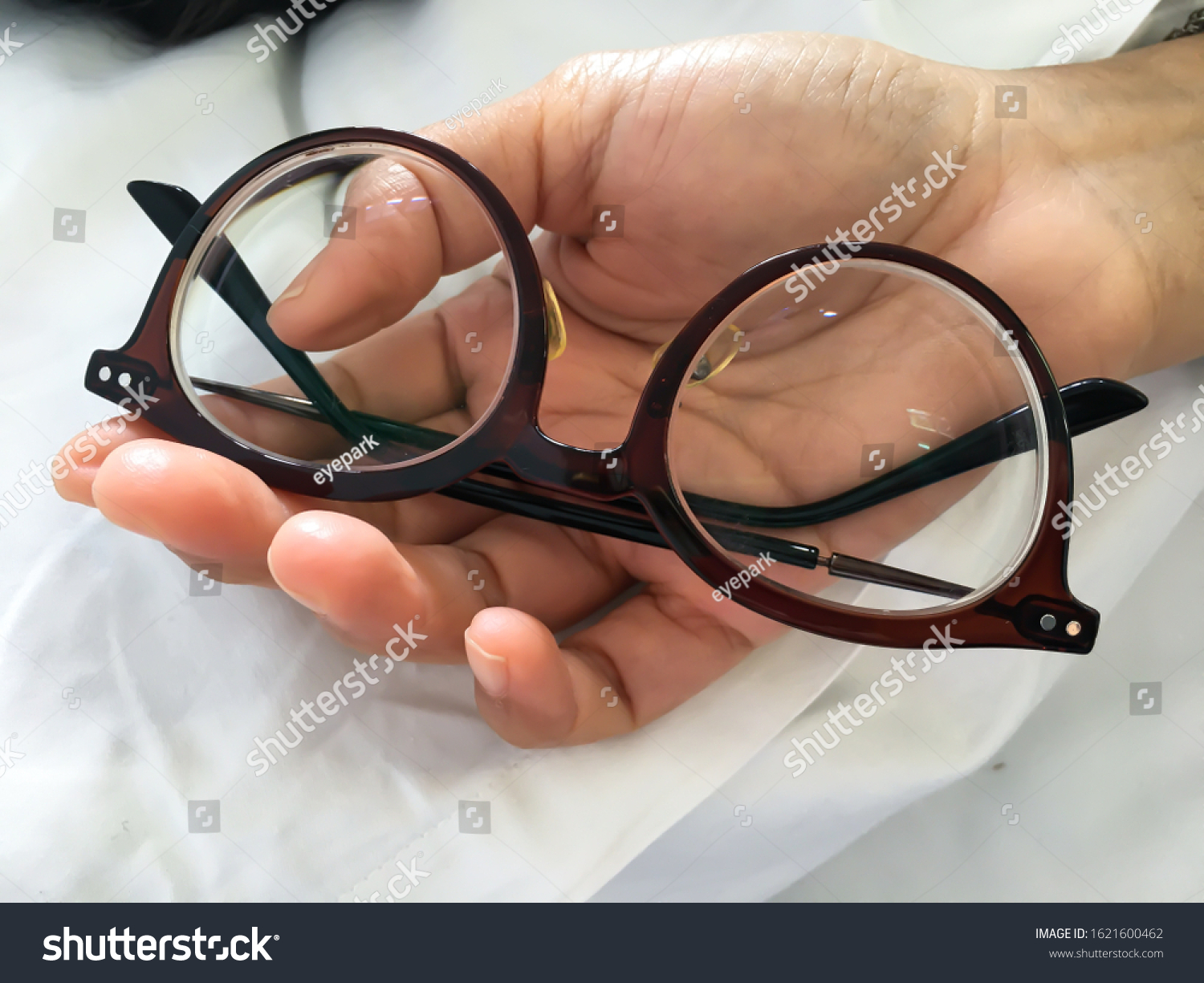 Woman's hand holding   shortsighted or nearsighted eyeglasses on white pillow, white bed background, Close up & Macro shot, Selective focus, Optical concept #1621600462
