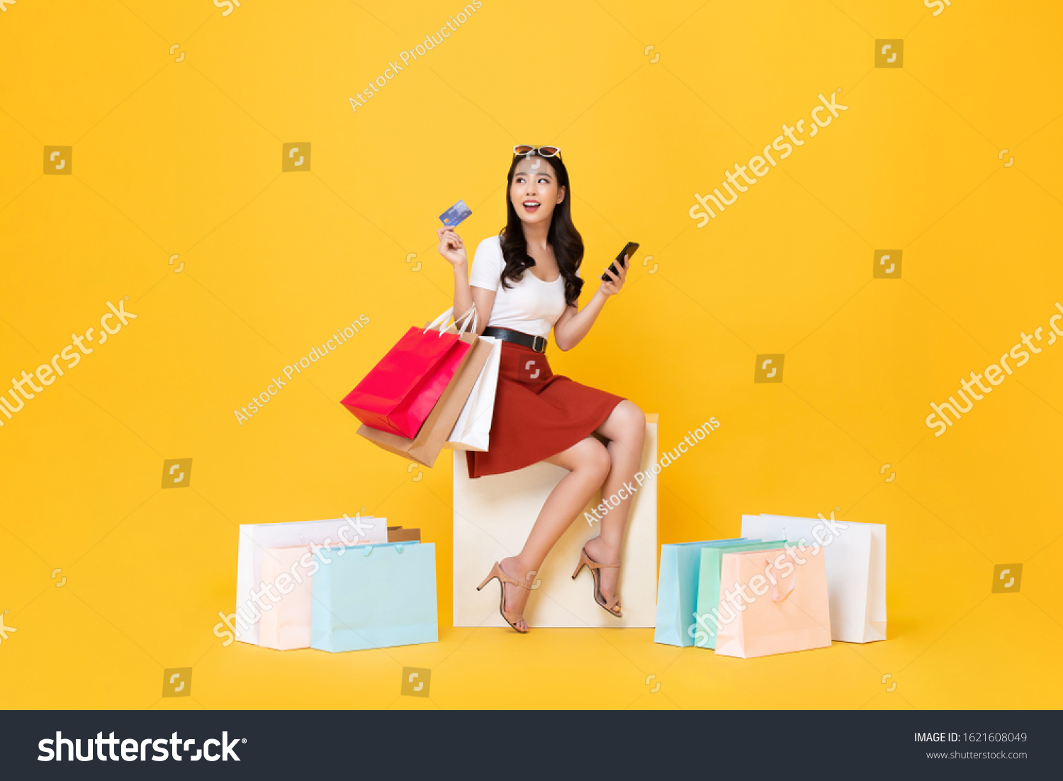 Beautiful Asian woman sitting and carrying shopping bags with credit card and mobile phone in hands on yellow background #1621608049