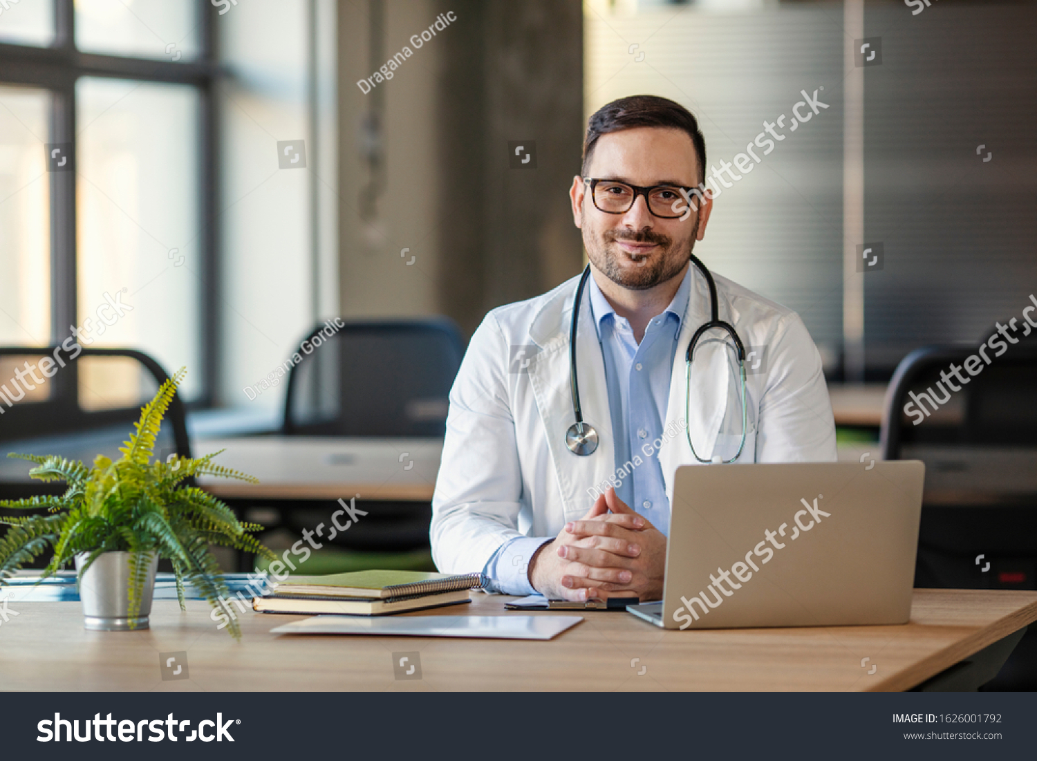 Doctor man sitting at the desk at his working place and smiling at camera. Perfect medical service in clinic. Portrait of a handsome young doctor. Happy doctor looking at camera  #1626001792