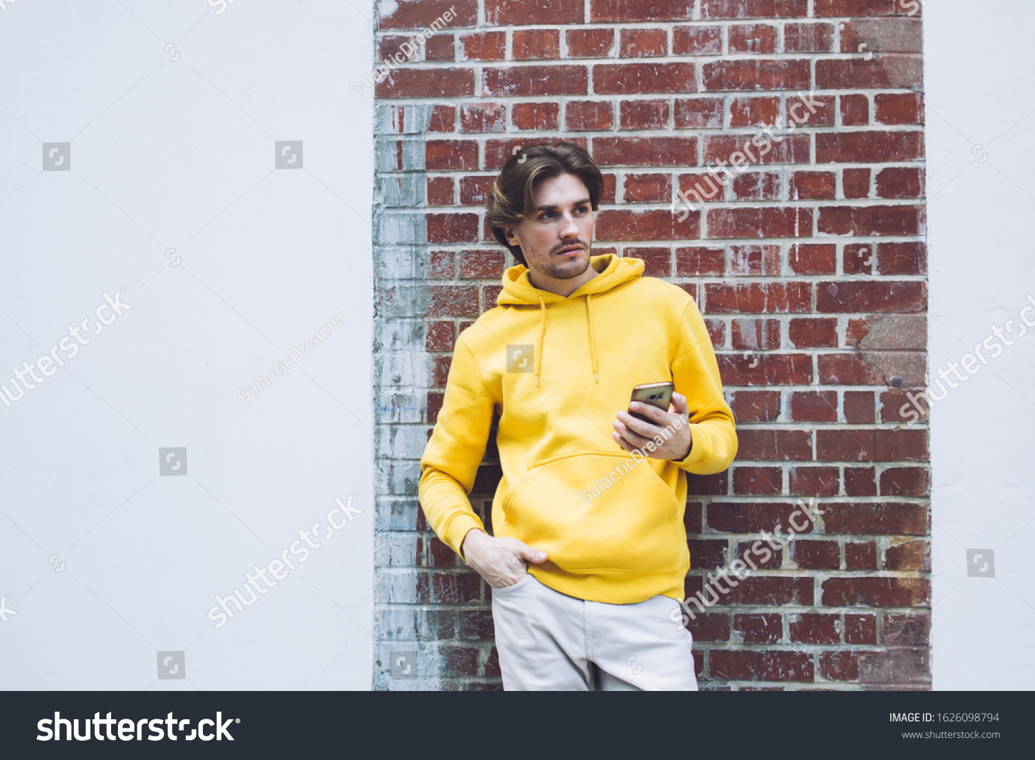 Pensive casually dressed young man in yellow sweatshirt standing back to brick wall with one hand in pocket while holding phone in other hand #1626098794