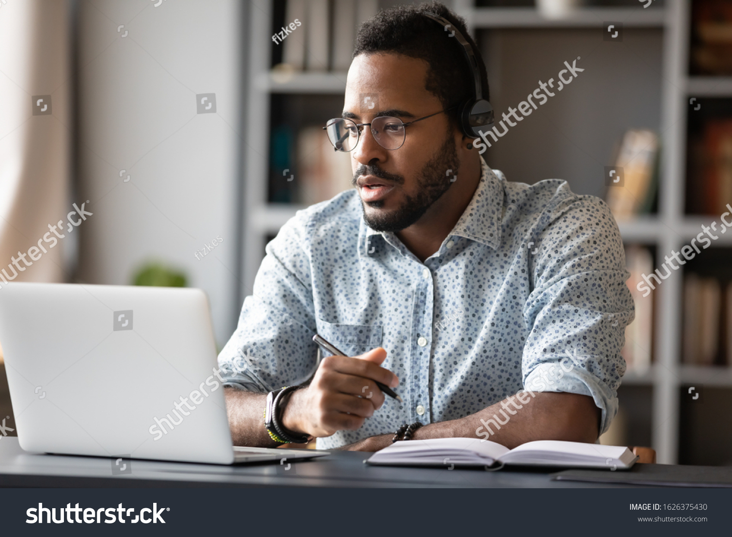 Focused young african businessman wear headphones study online watching webinar podcast on laptop listening learning education course conference calling make notes sit at work desk, elearning concept #1626375430