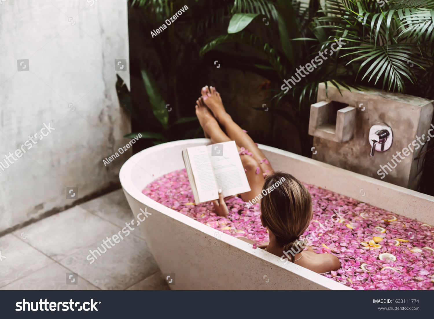 Woman reading book while relaxing in bath tub with flower petals. Organic spa relaxation in luxury Bali outdoor bath. #1633111774
