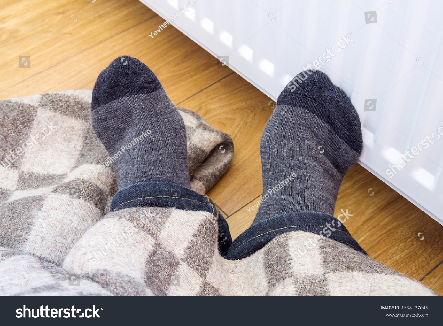 A person warms his legs under a woolen warm blanket near a home heater in cold winter time. The symbolic image of the heating season at home.  #1638127045