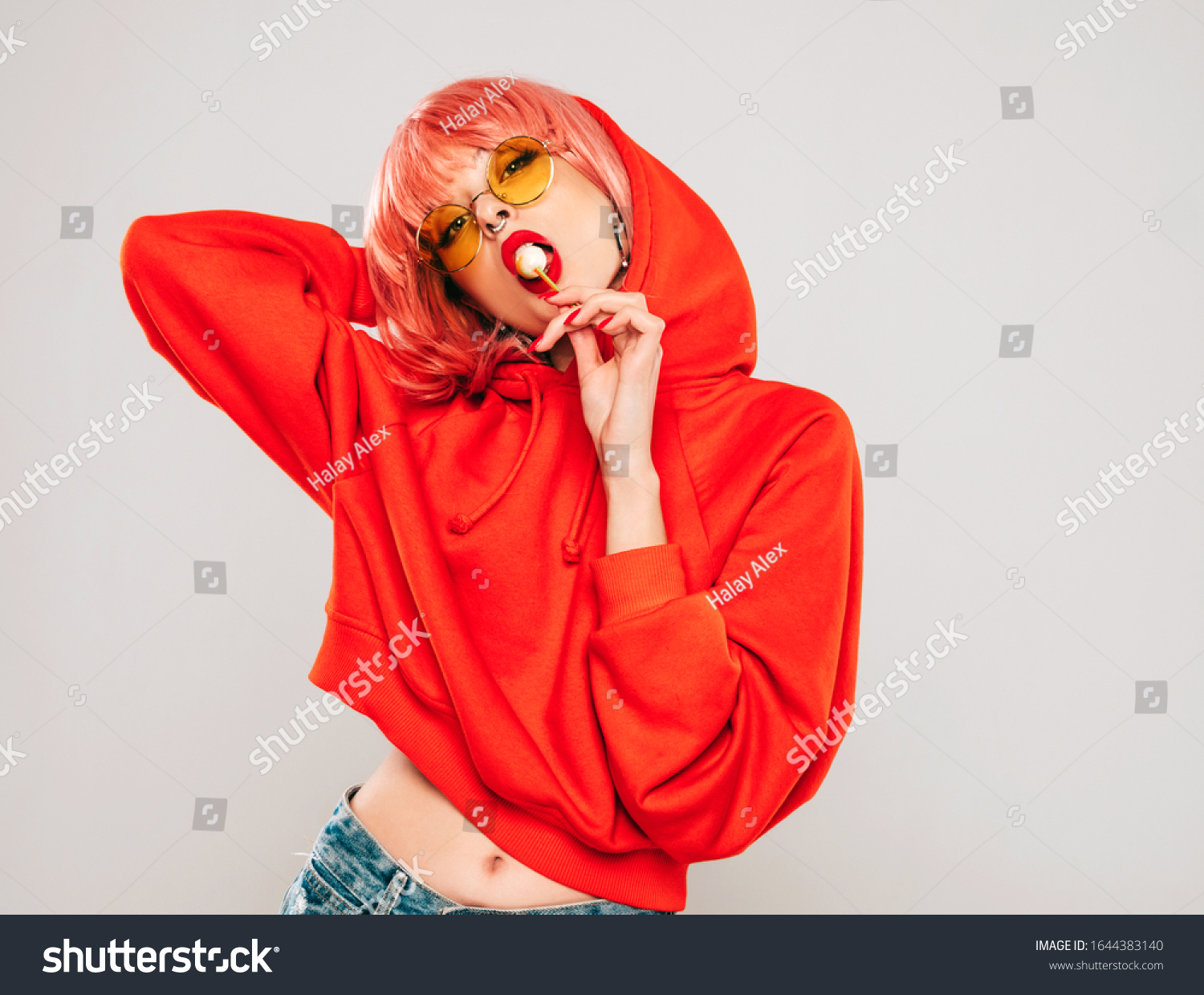 Young beautiful hipster bad girl in trendy red summer red hoodie and earring in her nose.Sexy carefree woman posing in studio on gray background in wig.Hot model licking round sugar candy #1644383140