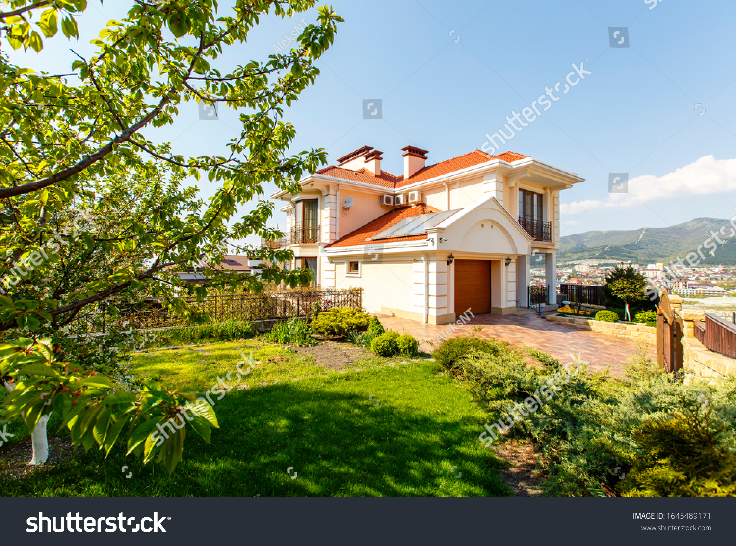 The facade of a classic Mediterranean two-storey cottage in the spring. The cottage is white with a red tiled roof. In front of the cottage there is a green lawn and flowering trees.  #1645489171