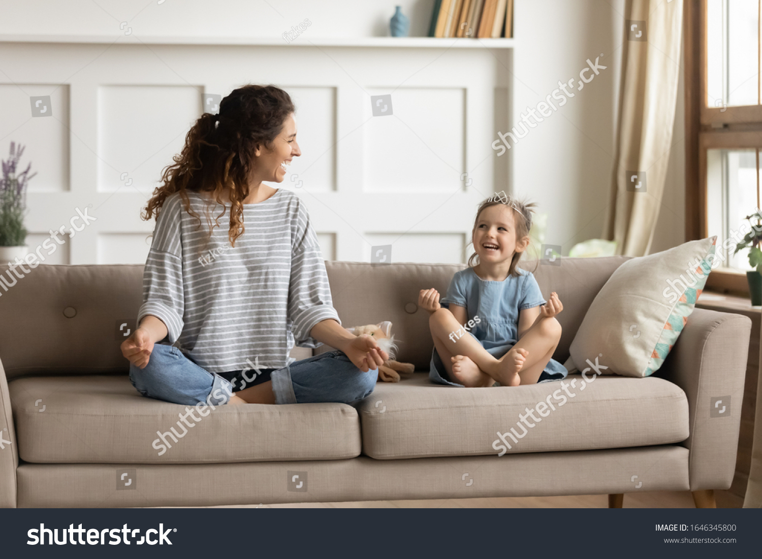 Full length front view smiling young curly mother sitting on comfortable couch with cute playful little preschool daughter in lotus position. Happy mommy practicing yoga exercised with small child. #1646345800