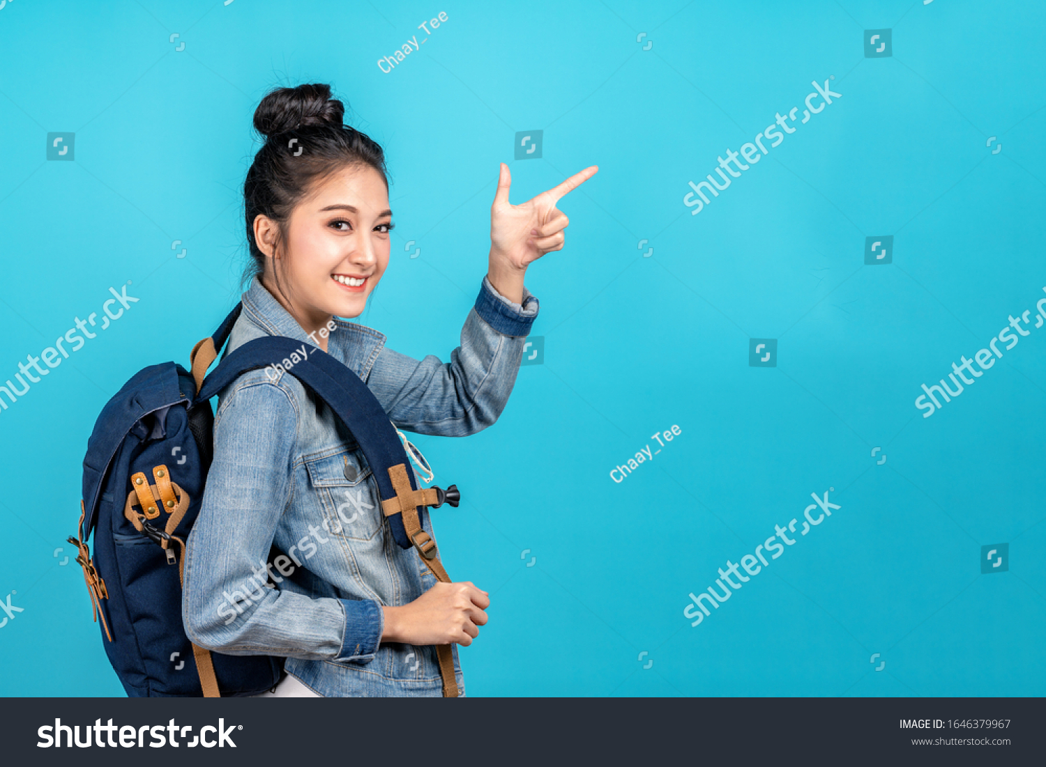 Happy asian woman travel backpacker standing pointing hands to copyspace on blue background. Cute asia girl smiling wearing casual jeans shirt and finger pointing to aside for present promotions. #1646379967
