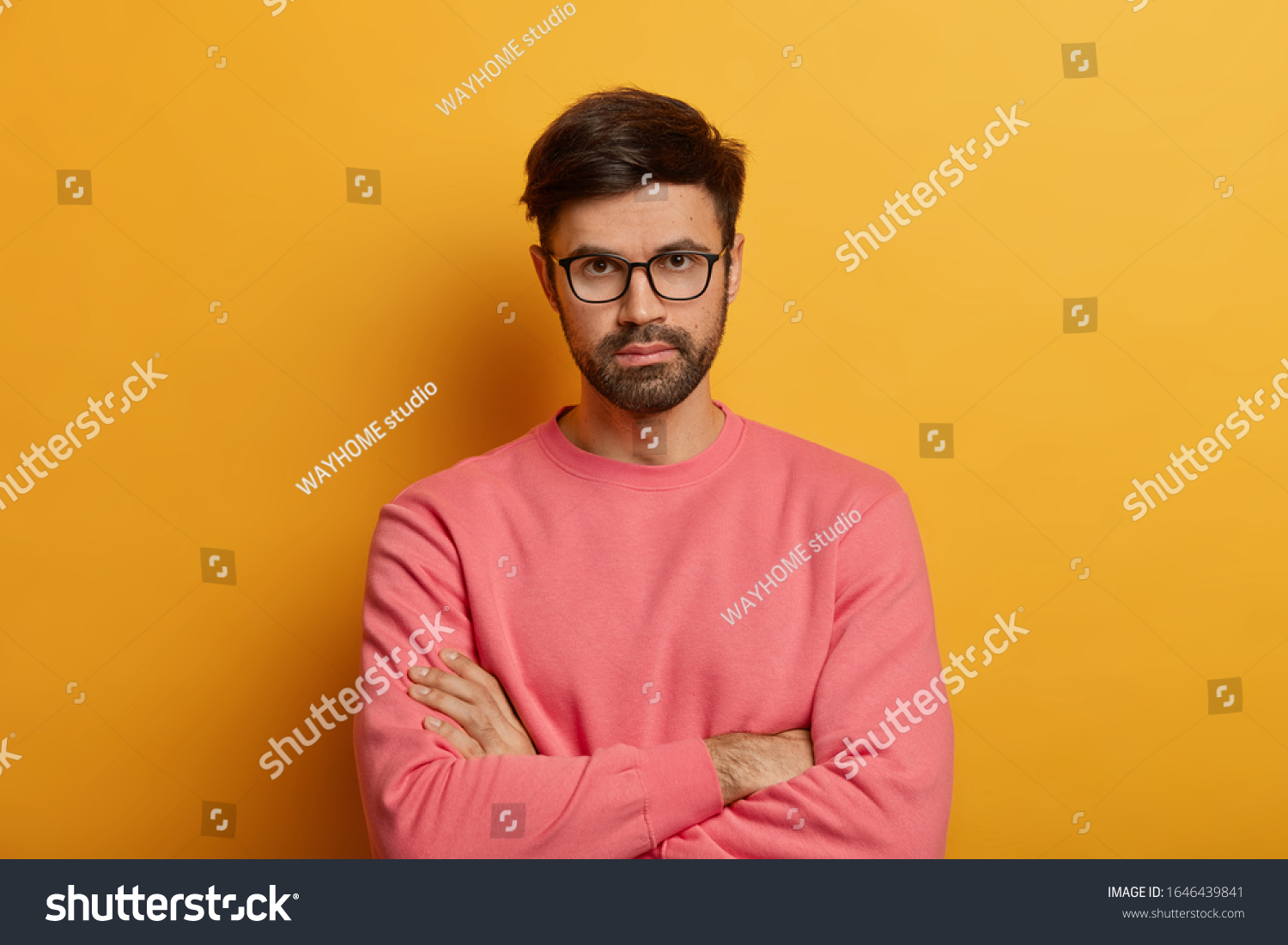 Portrait of self confident serious male entrepreneur keeps hands crossed, looks seriously directly at camera, stands against yellow background discusses necessary things with partner wears pink jumper #1646439841