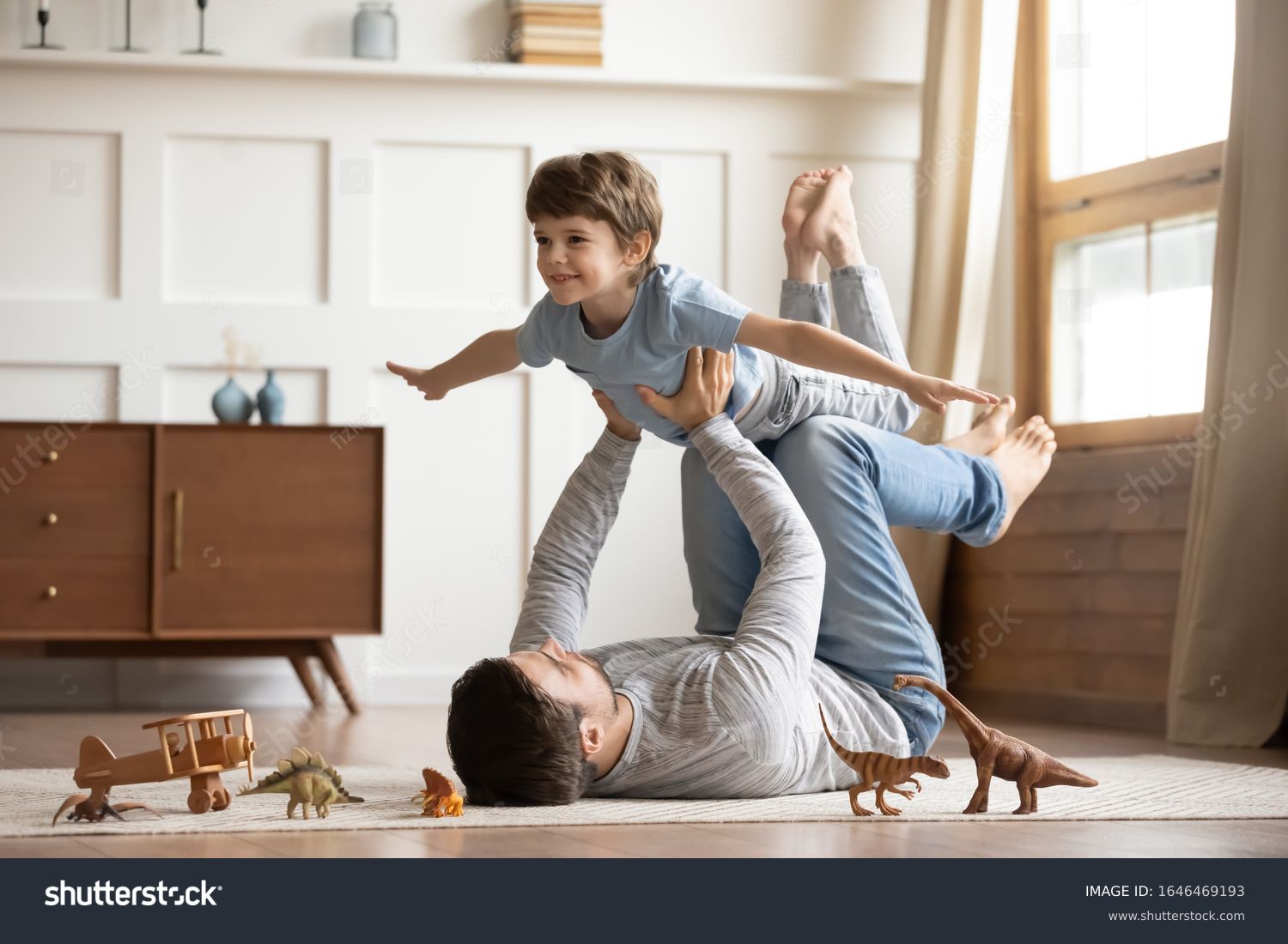 Joyful young man father lying on carpet floor, lifting excited happy little child son at home. Full length carefree two generations family having fun, practicing acroyoga in pair in living room. #1646469193