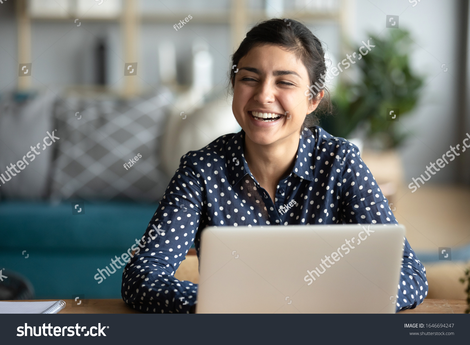 Overjoyed young Indian woman sit at desk in living room talk with friend, happy millennial ethnic girl distracted from computer work laugh have fun engaged in pleasant conversation at home #1646694247