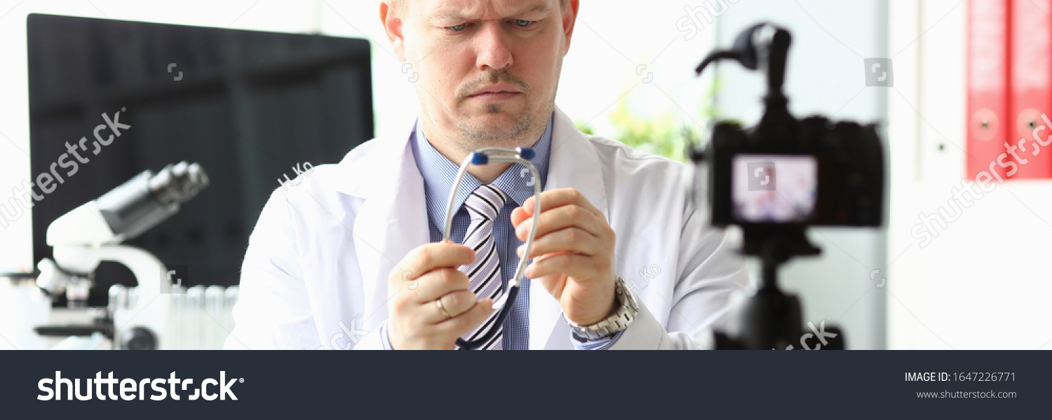 Portrait of serious blogger looking attentively at stethoscope. Handsome doctor recording video for vlog in modern office for internet channel. Blogging concept #1647226771