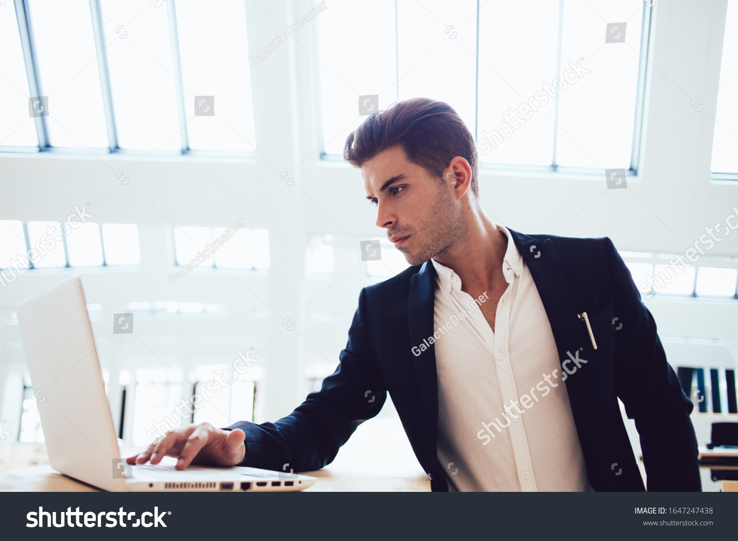 Concentrated youthful smart male freelancer focusing on laptop screen while typing and working on business project in light modern office #1647247438