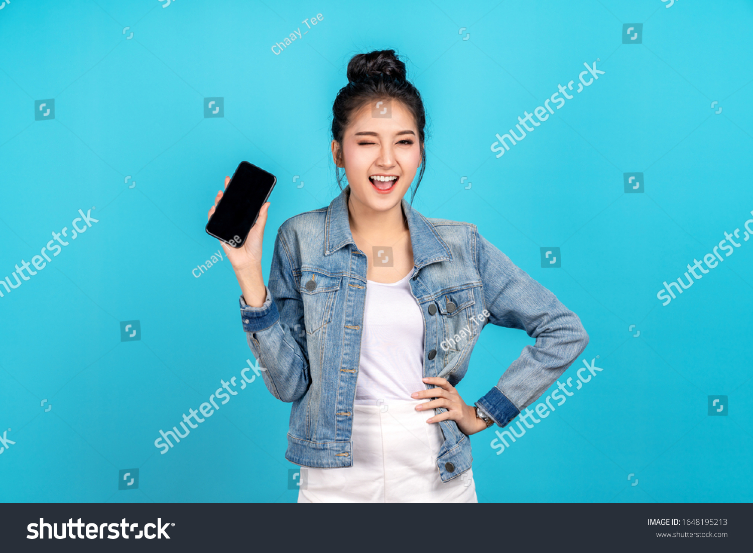 Happy asian woman feeling happiness, blinks eyes and standing hold smartphone on blue background. Cute asia girl smiling wearing casual jeans shirt and connect internet shopping online and present #1648195213