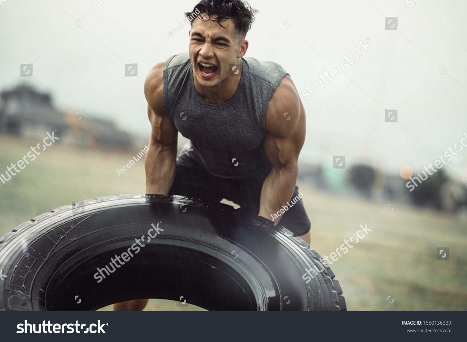 Tough young male athlete doing a tire flip exercise in the rain. Muscular man doing cross training outdoors on field. #1650136339