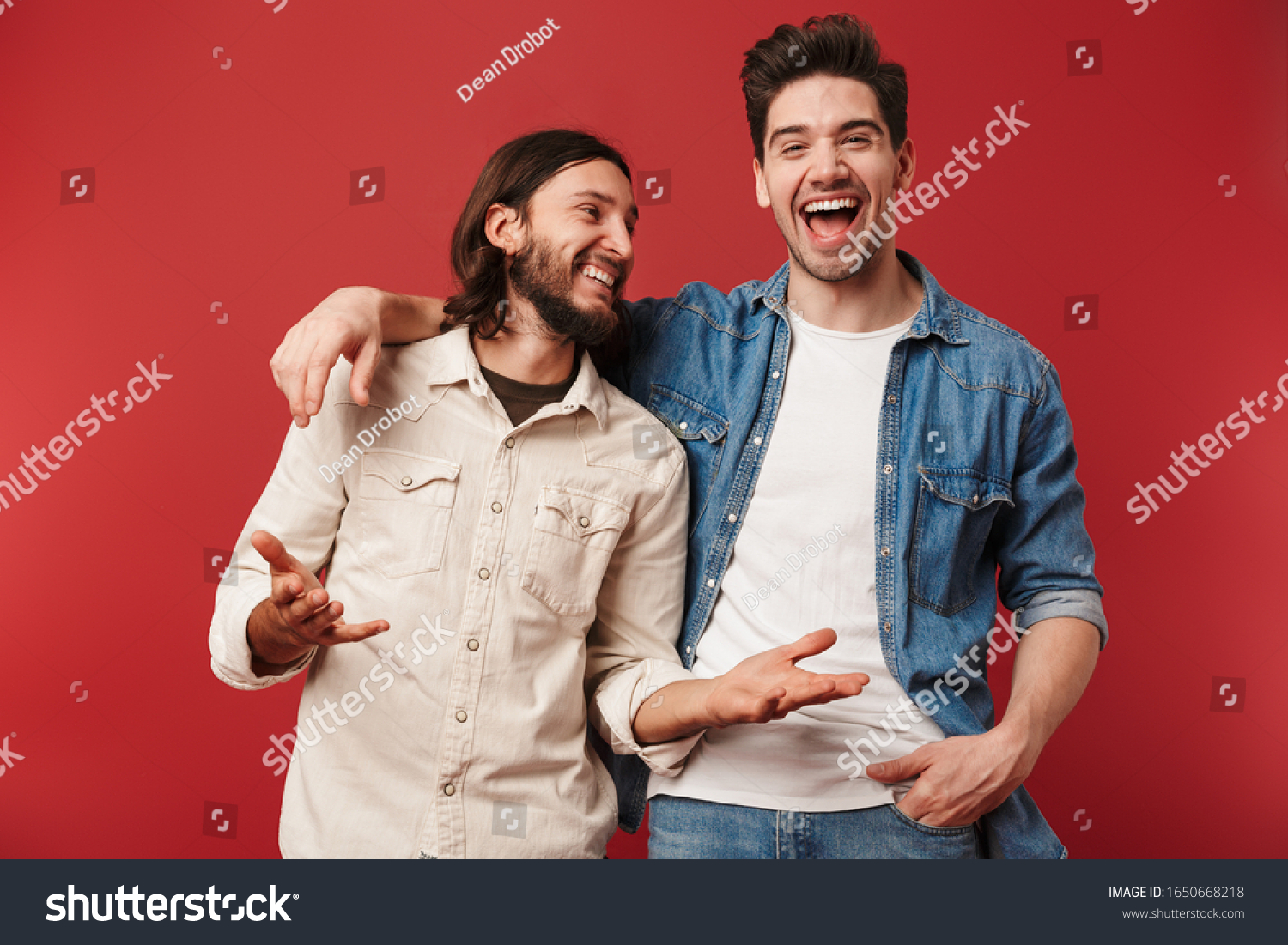 Two cheerful young men wearing casual clothes standing isolated over red background, laughing #1650668218