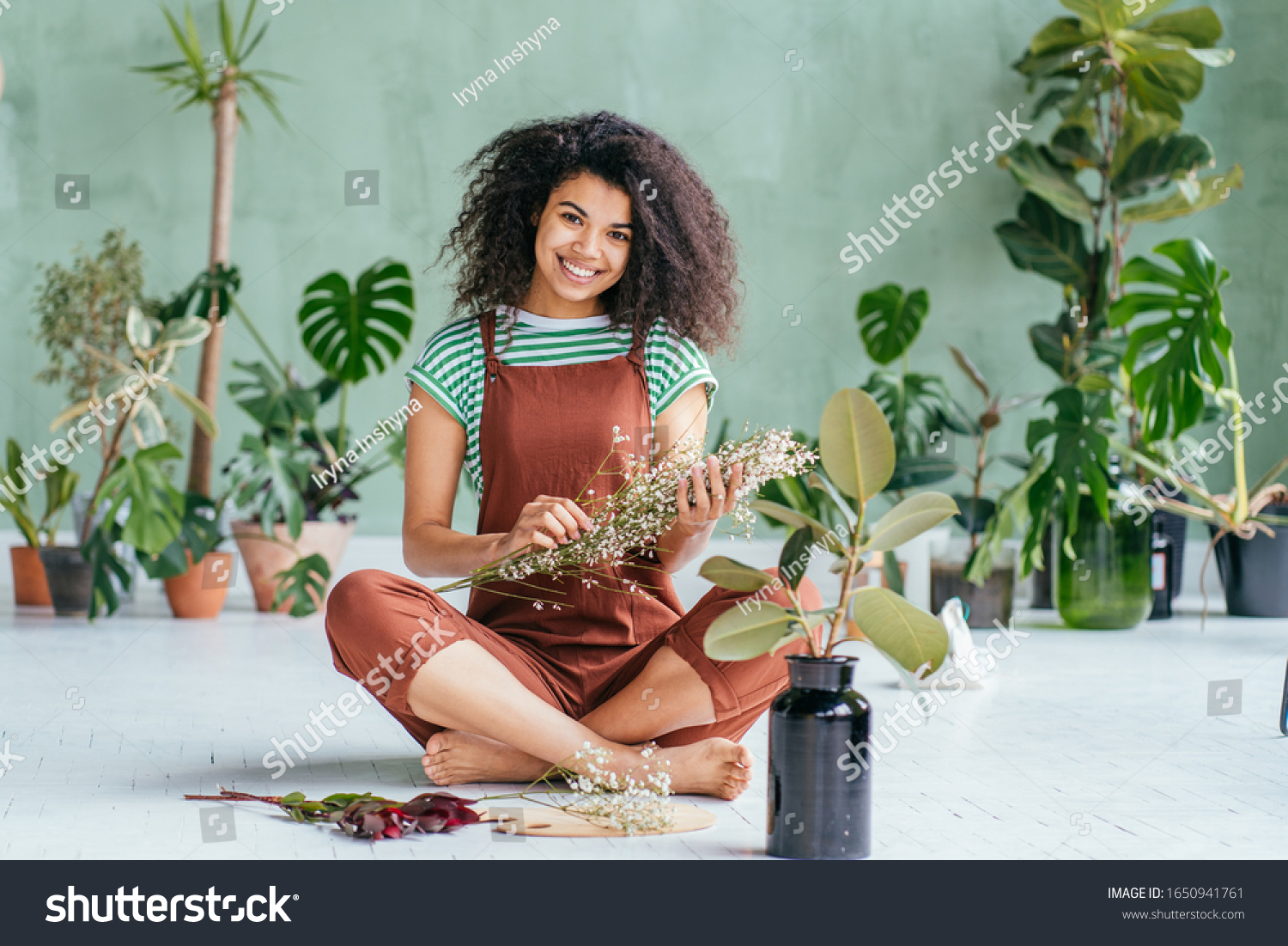 Young woman cultivating home plants.Small business.Sensual mixed race female florist with flowers in hands against background of indoor plants. Life lover, zero waste, inspiration, summer mood concept #1650941761