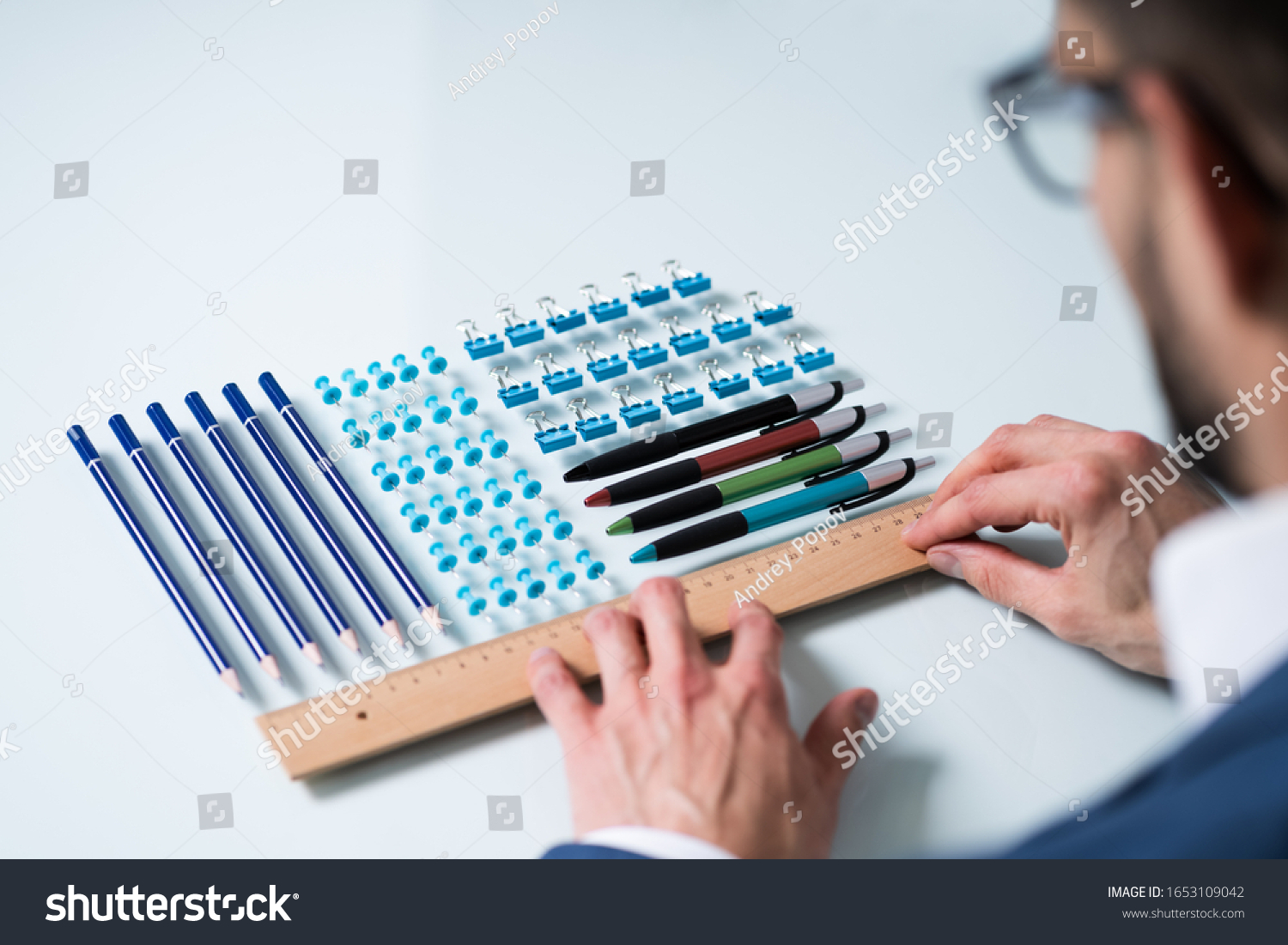 A Person's Hand Arranging Pencils And Multi Colored Pushpins In A Row On White Background #1653109042