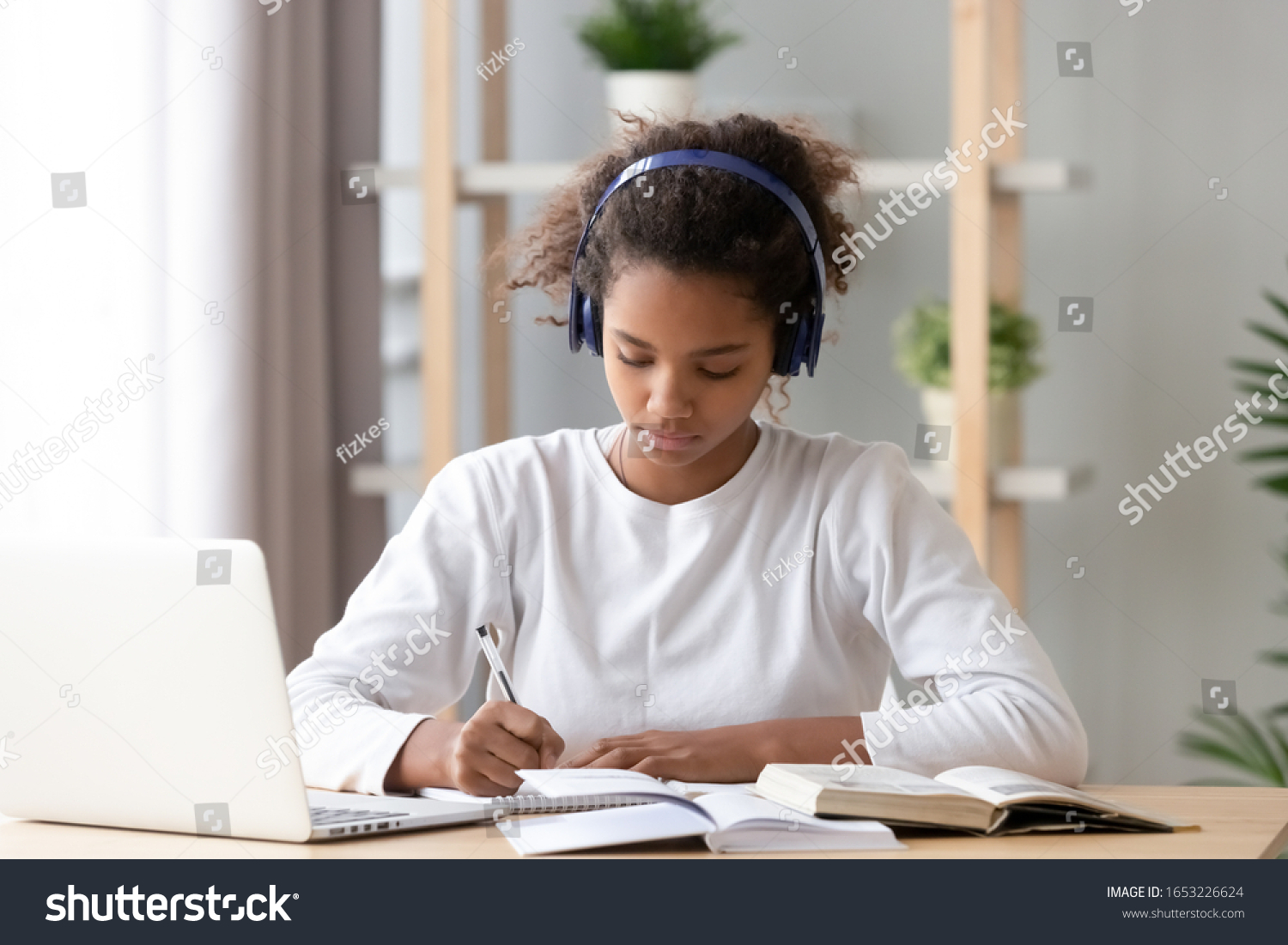 Focused african american teenage girl wearing headphones writing notes study with laptop and books, serious black female high school teen student listening audio course or music while doing homework #1653226624
