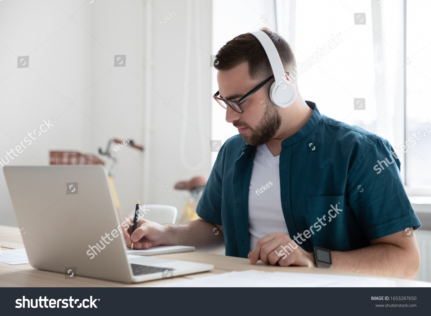 Focused young man businessman company worker employee in glasses wearing wireless headphones, watching educational webinar lecture seminar on laptop online, writing down notes in modern office. #1653287650